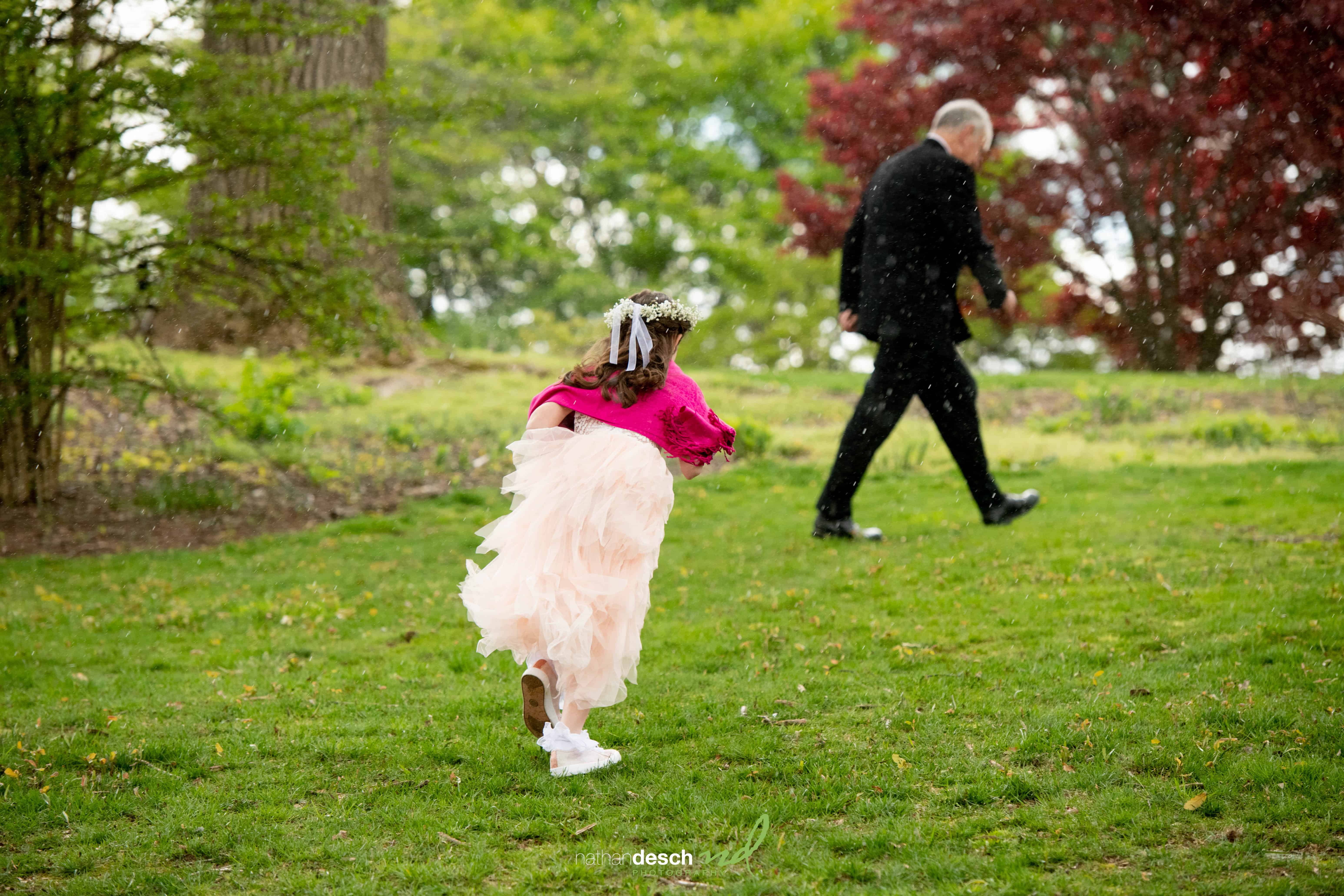 Young flower girl in pink dress runs on grass at Frelinghuysen Arboretum wedding, chasing man in suit with trees behind.