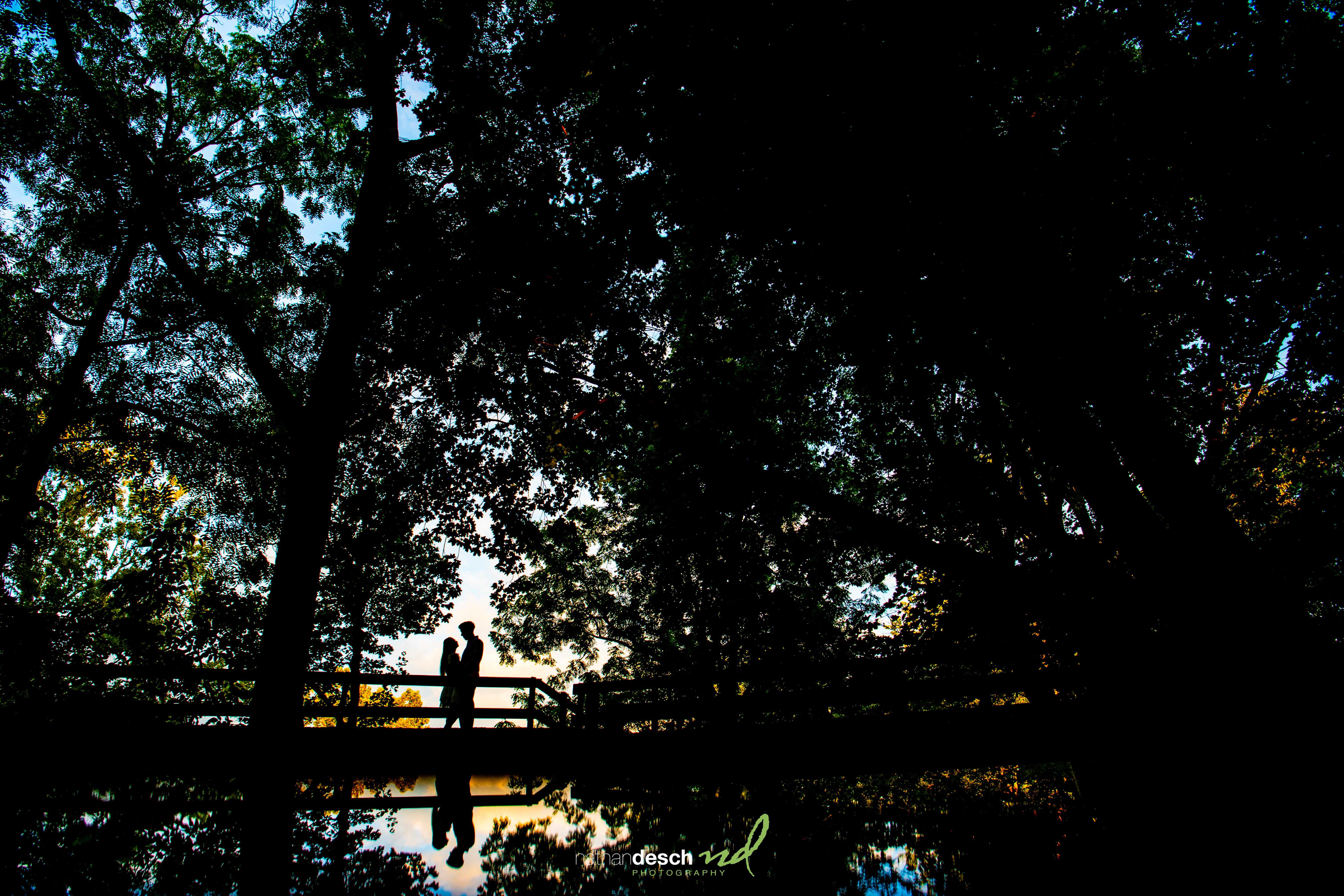 Silhouette of a couple embracing on a wooden bridge at Lock Ridge Park at sunset, reflected in the water below surrounded by trees.