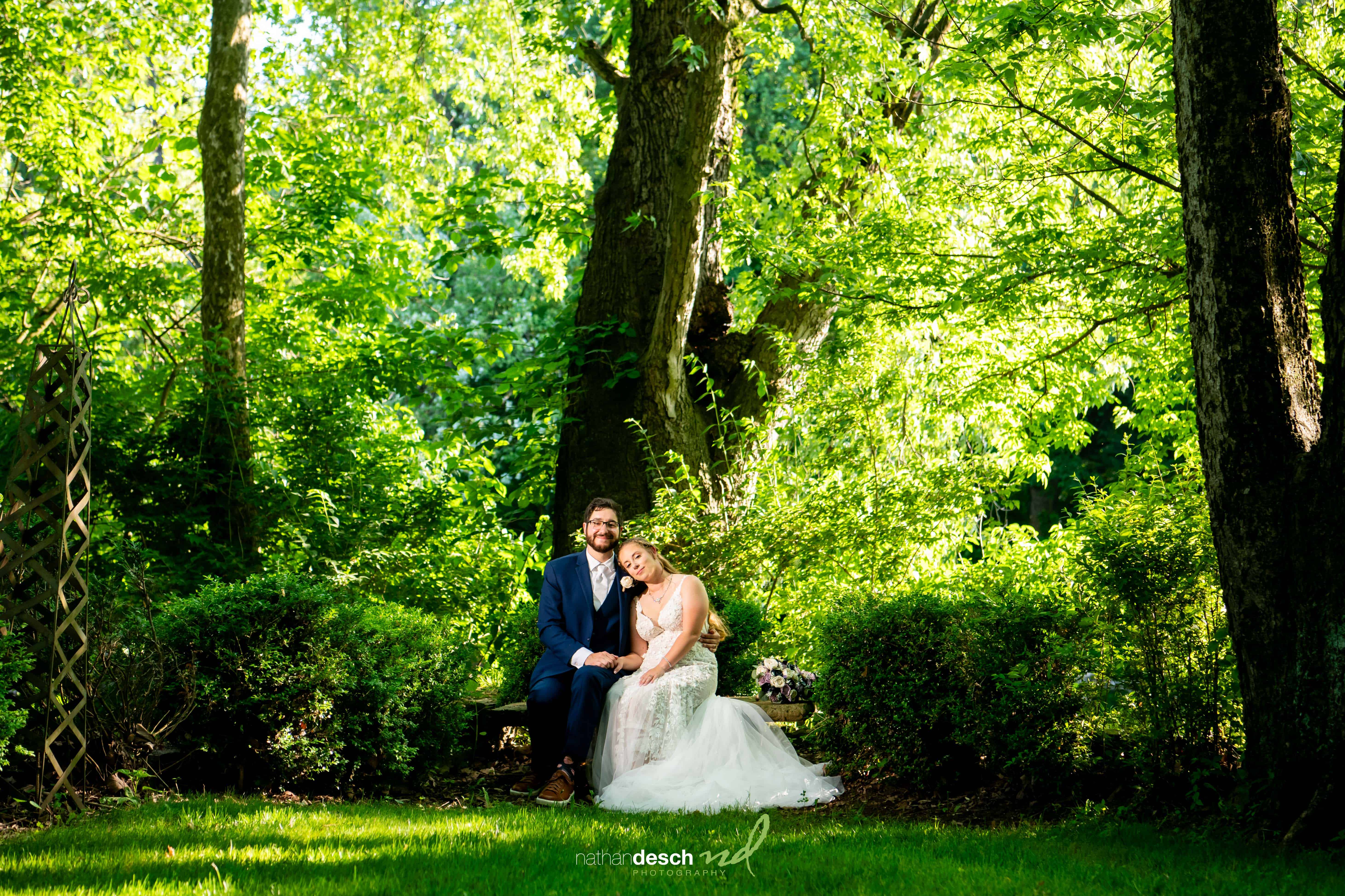 Bride and groom share a quiet moment on a garden bench at Riverdale Manor spring wedding, surrounded by lush greenery.