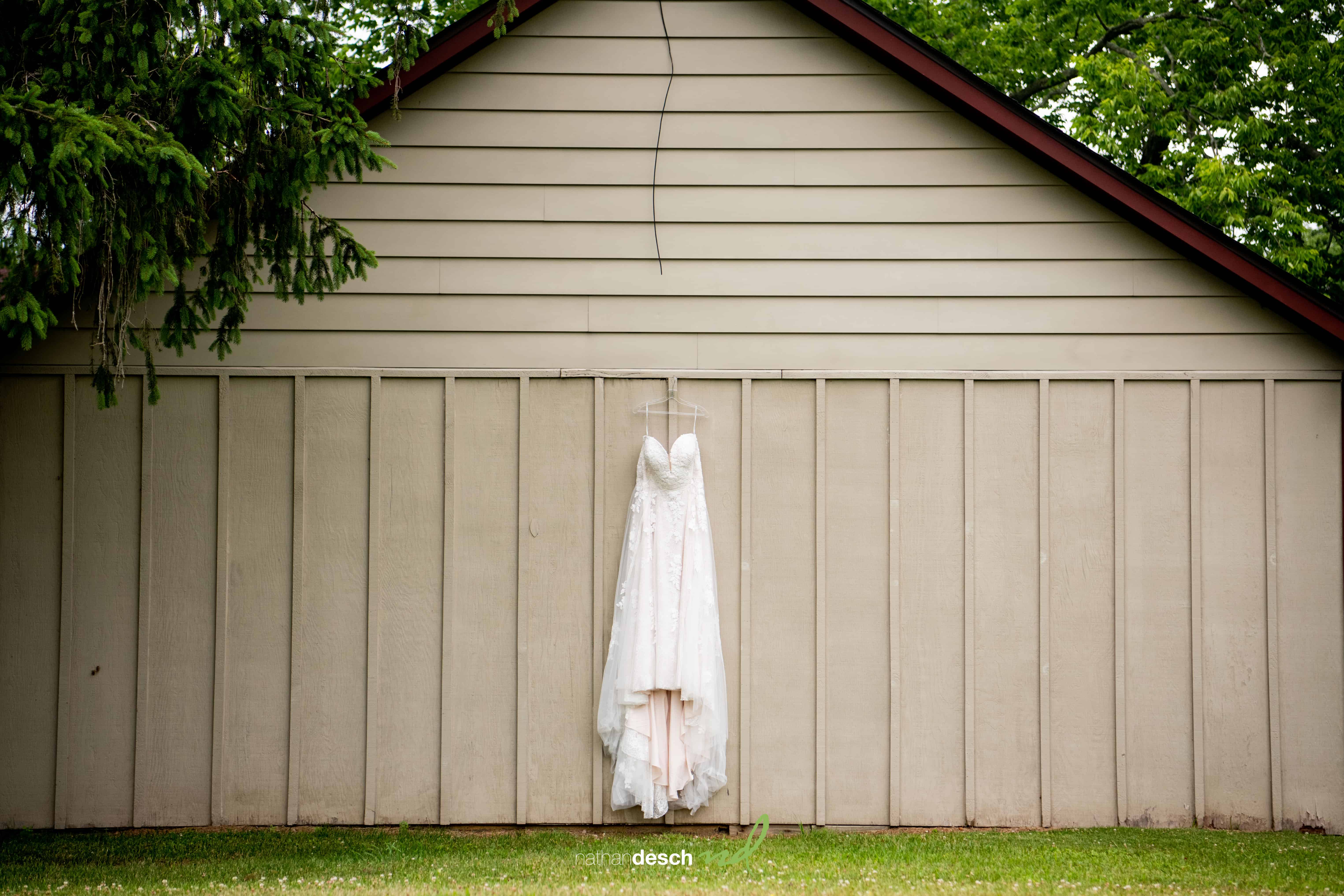 White strapless wedding dress on hanger against beige wood wall with green trees, awaiting bride before ceremony.