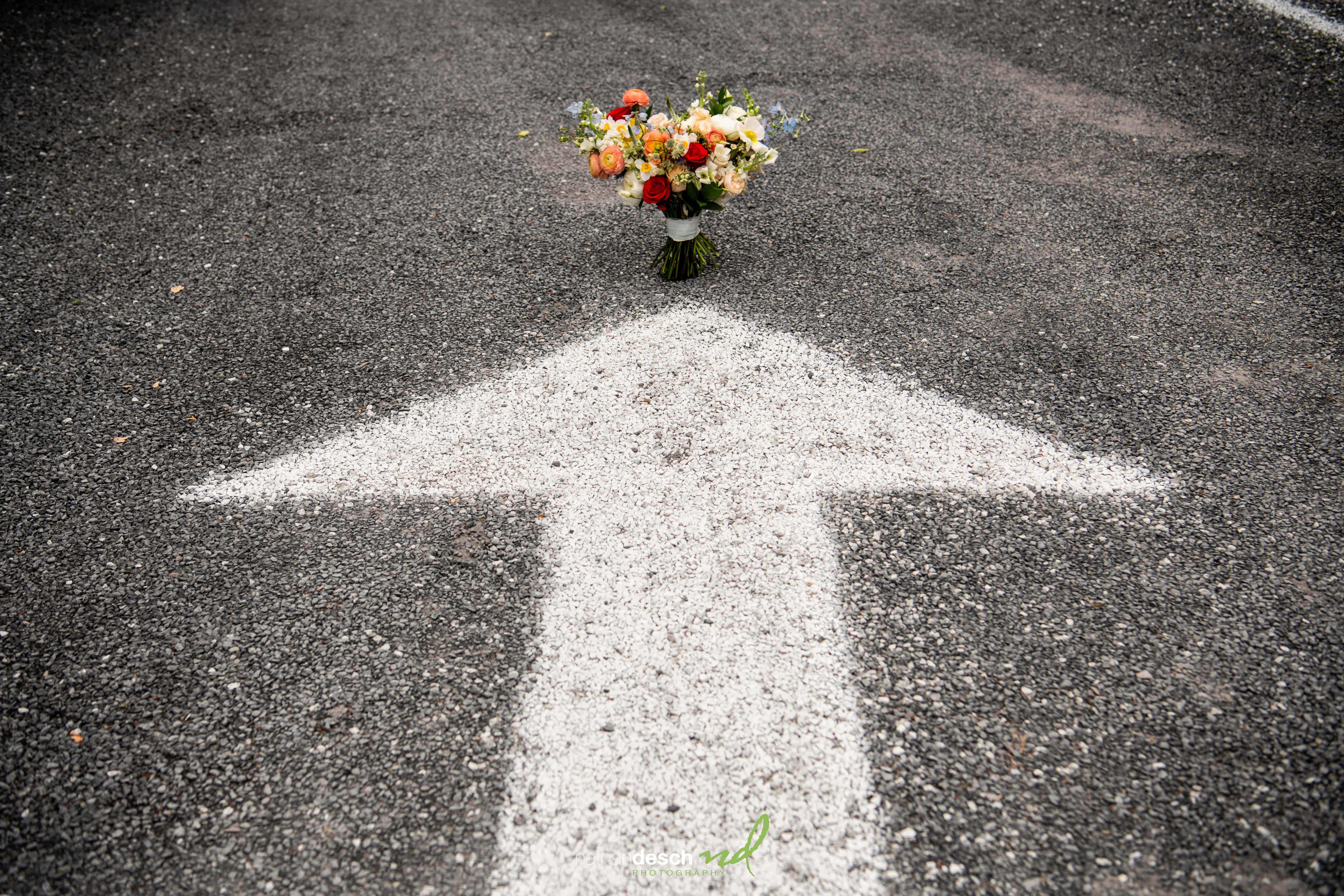 Bridal bouquet on asphalt road at arrow tip; unique wedding photo symbolizing Jake and Erika’s forward journey.