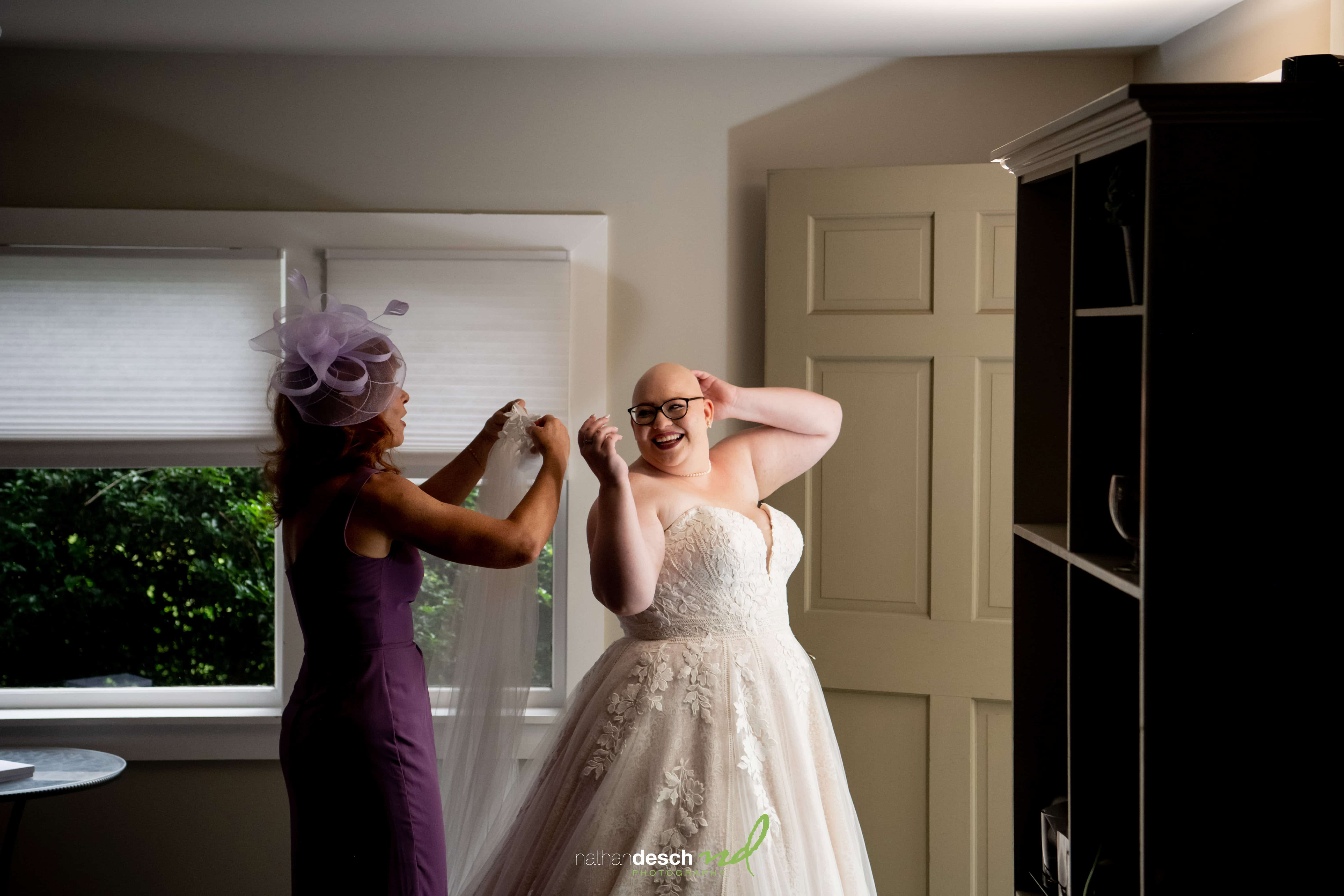 Bride in strapless white gown adjusting glasses as friends help with veil in softly lit room, wedding preparation moment.