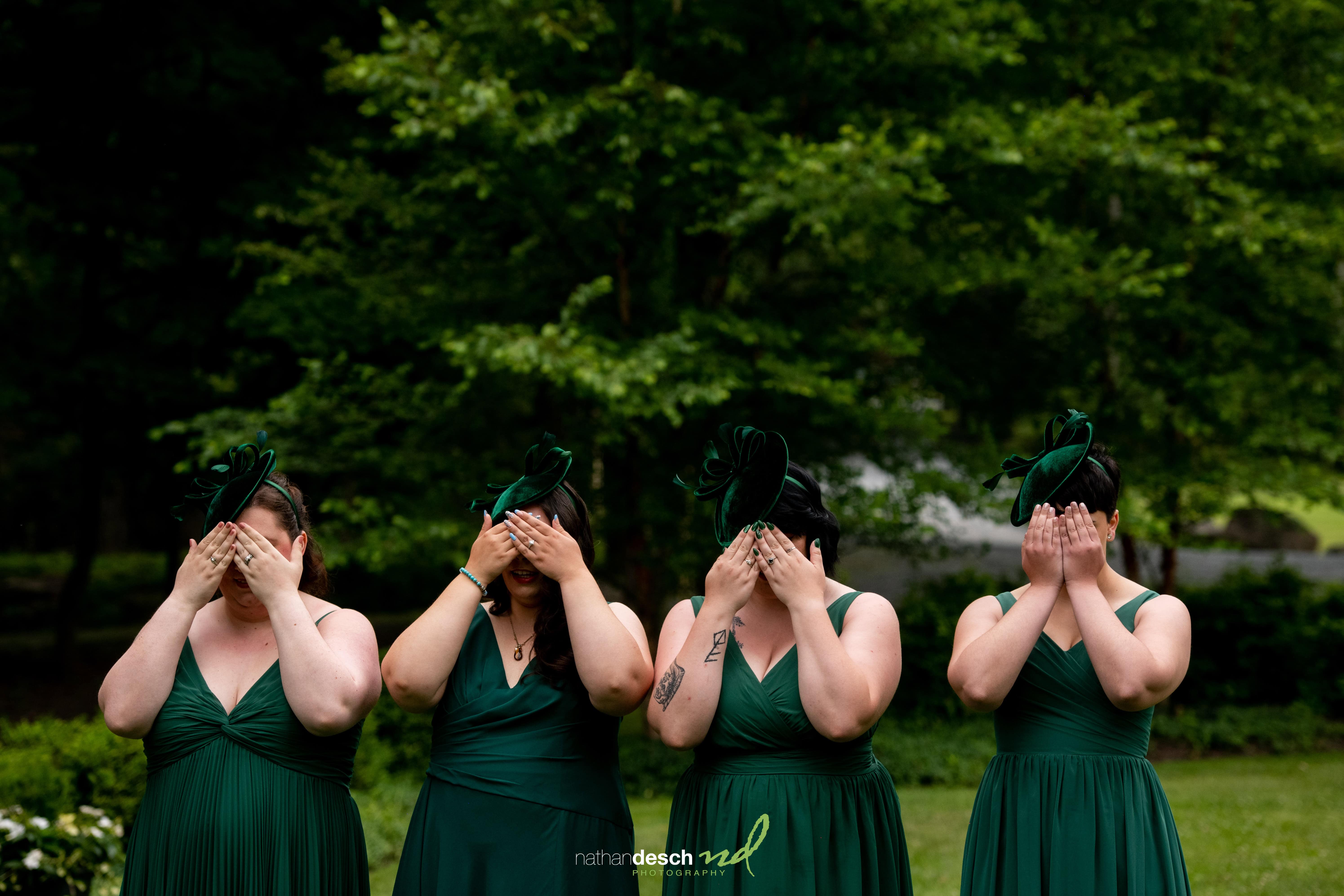 Bridesmaids in matching dark green dresses and headpieces cover their eyes outdoors with lush greenery in the background.