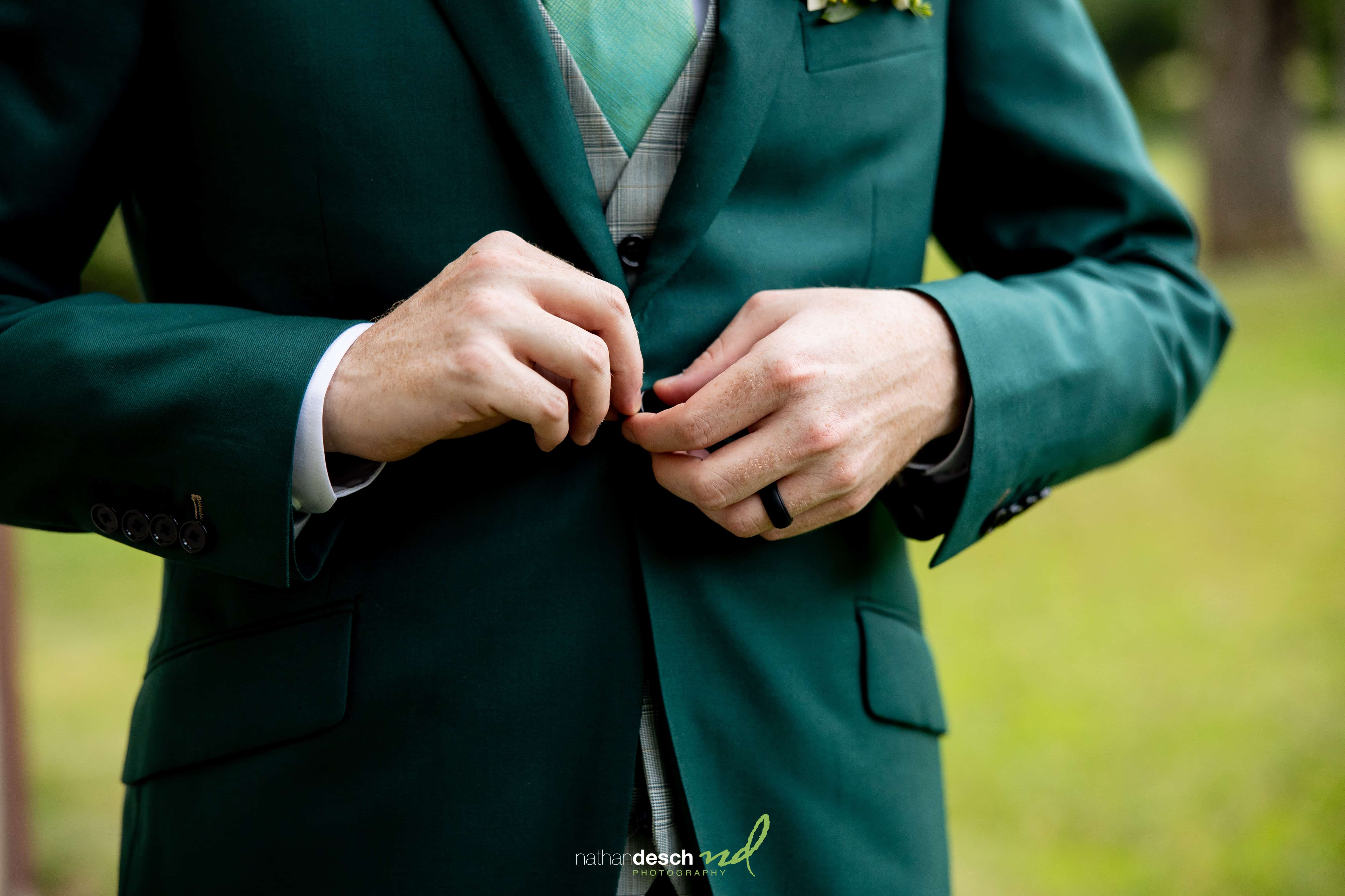 Groom in green suit and plaid tie buttons jacket outdoors, close-up of hands and torso on grassy wedding venue.