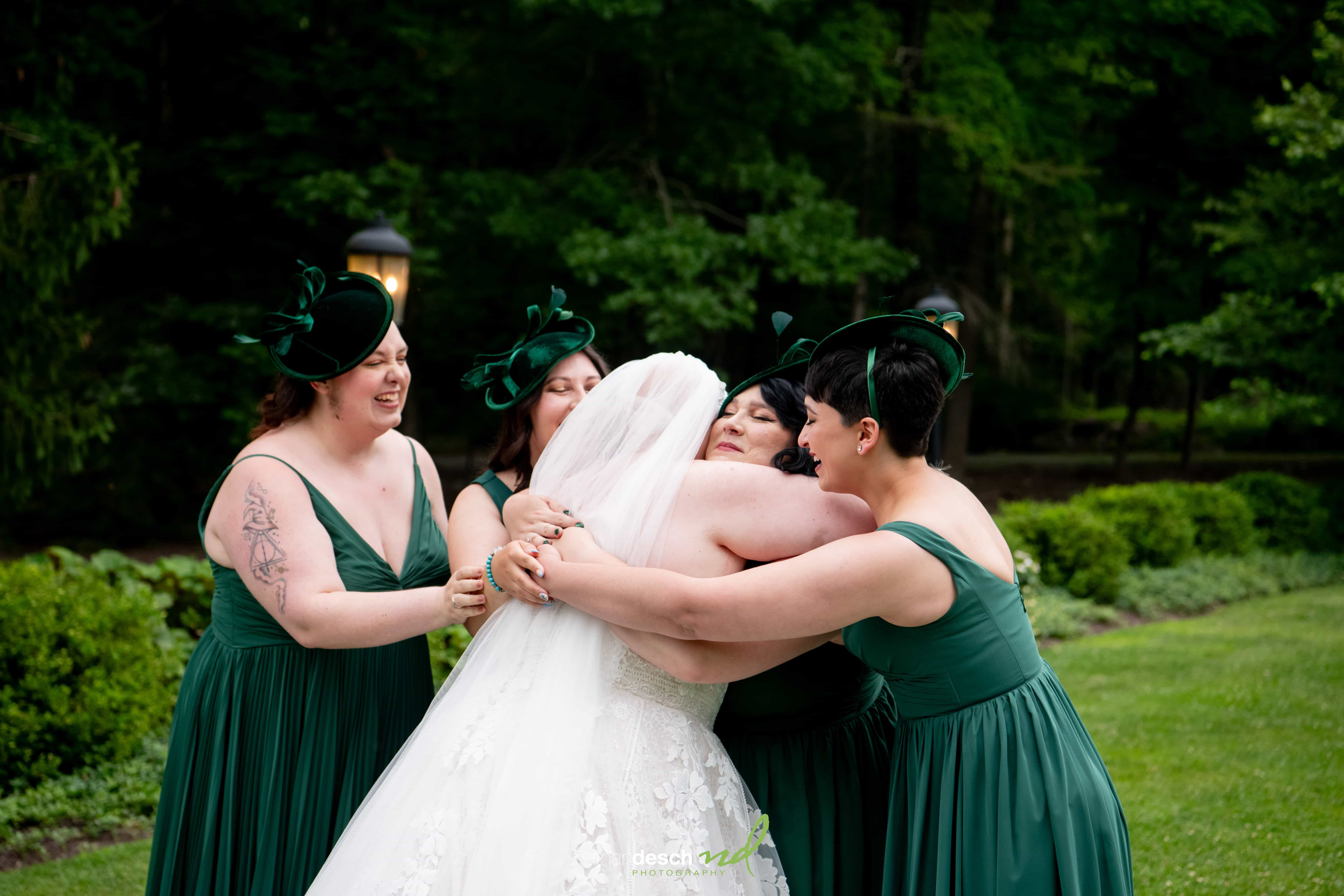 Bride in white dress hugs bridesmaids in dark green dresses and hats, smiling outdoors in a lush garden wedding scene.