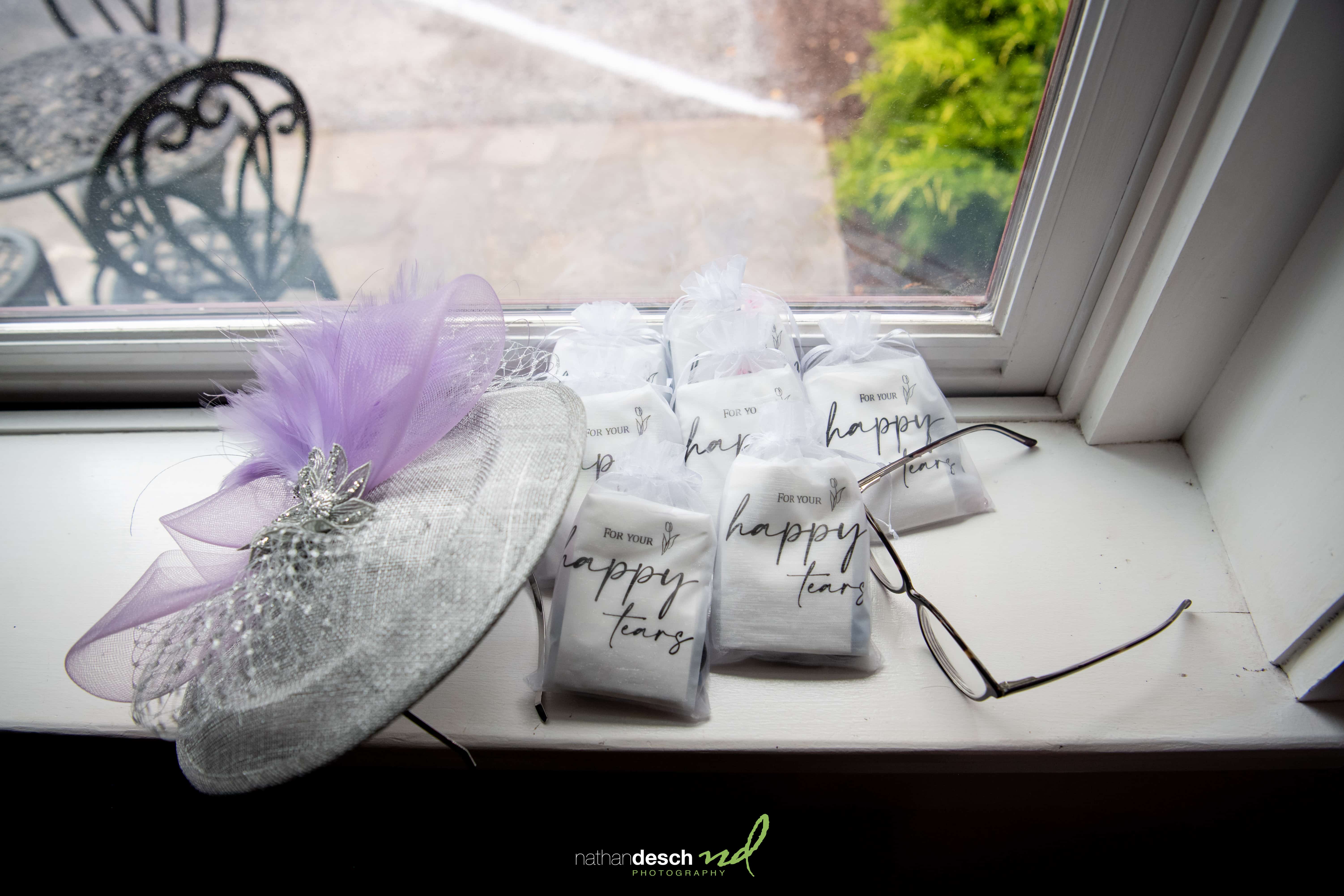 Decorative hat with purple tulle, glasses, and happy tears bags arranged on windowsill for wedding detail shot.