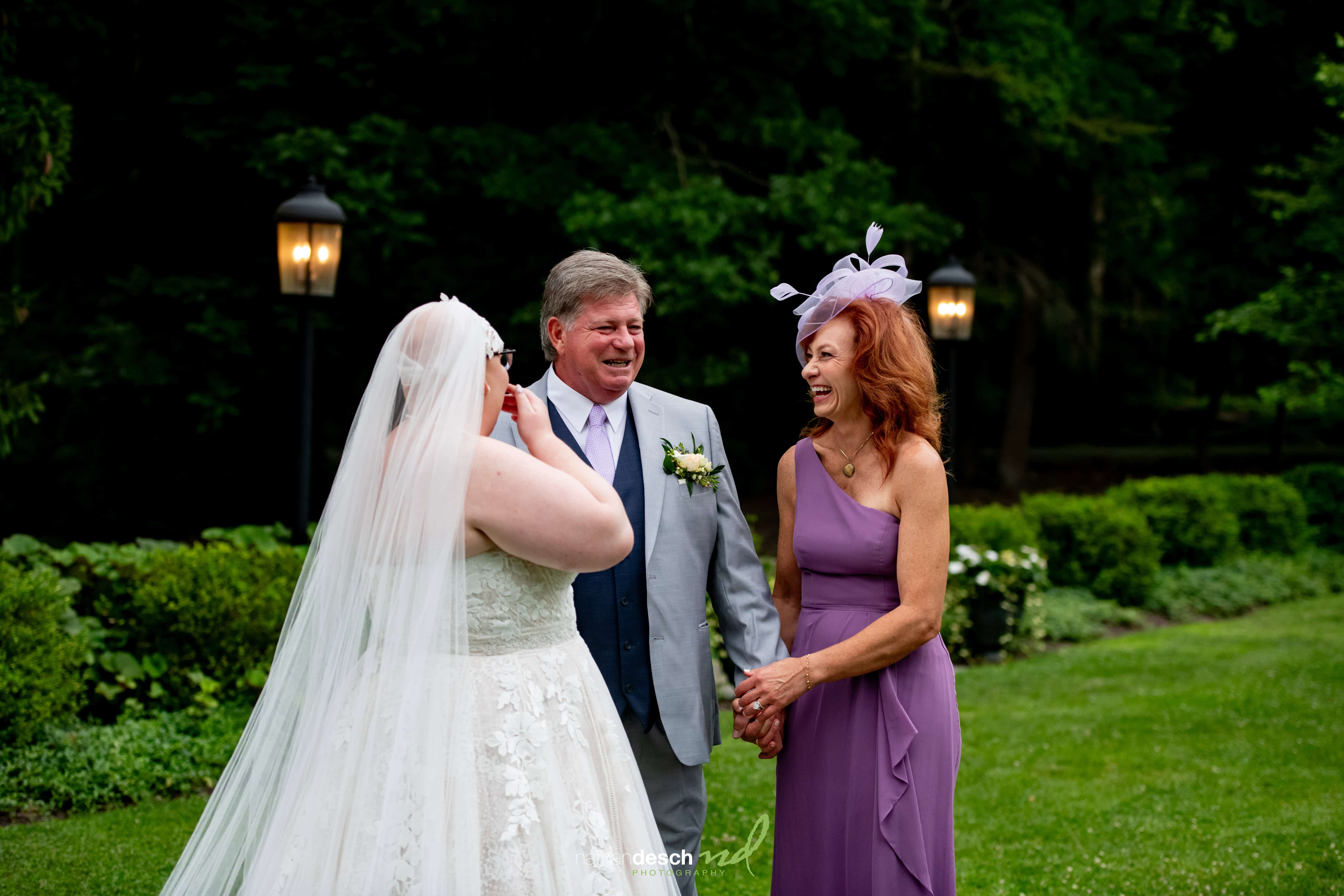 Bride in white dress wiping tears while smiling with parents in garden, surrounded by greenery and twinkling wedding lights.