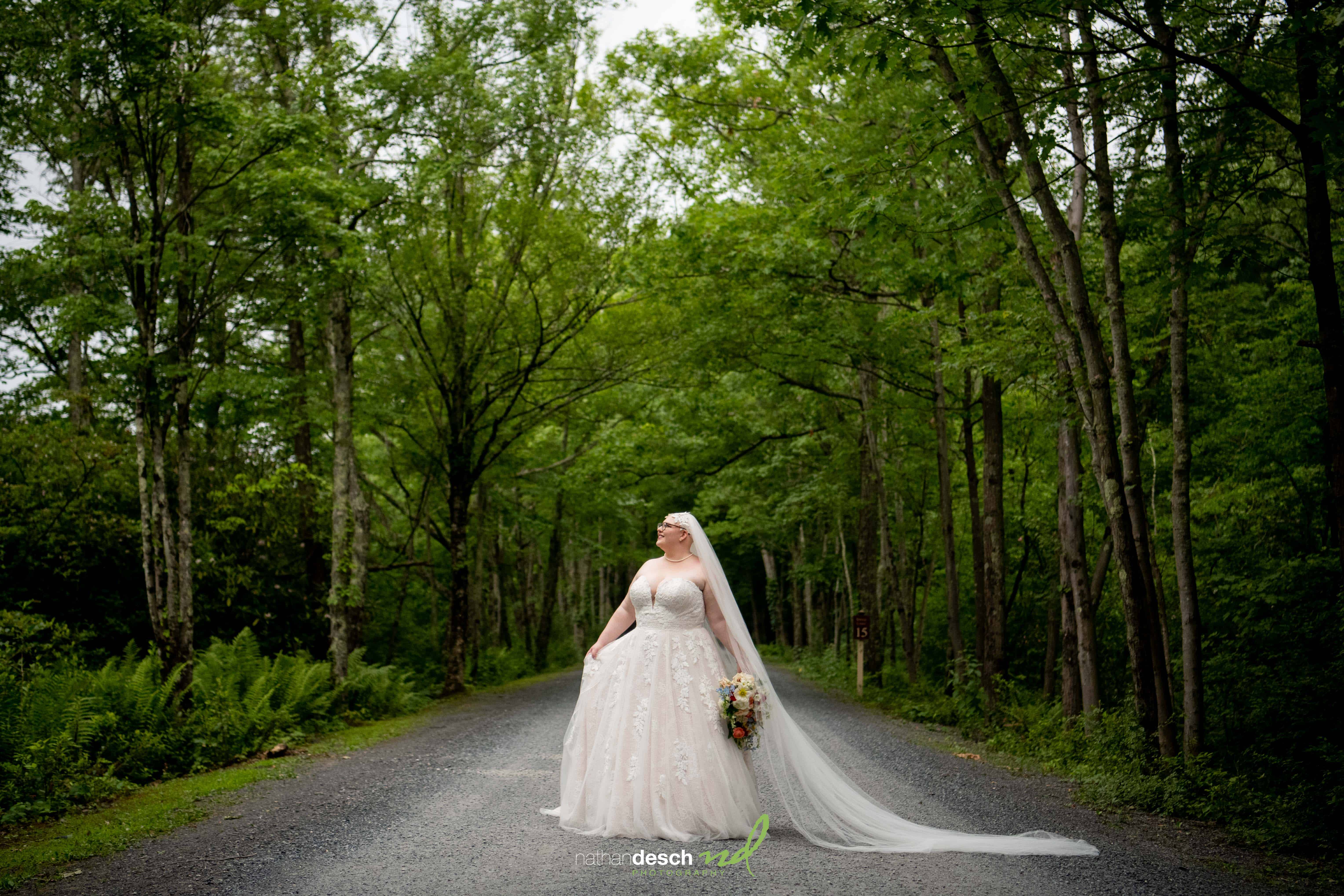 Bride in white gown and long veil stands on tree-lined road with bouquet, surrounded by lush greenery under cloudy sky.