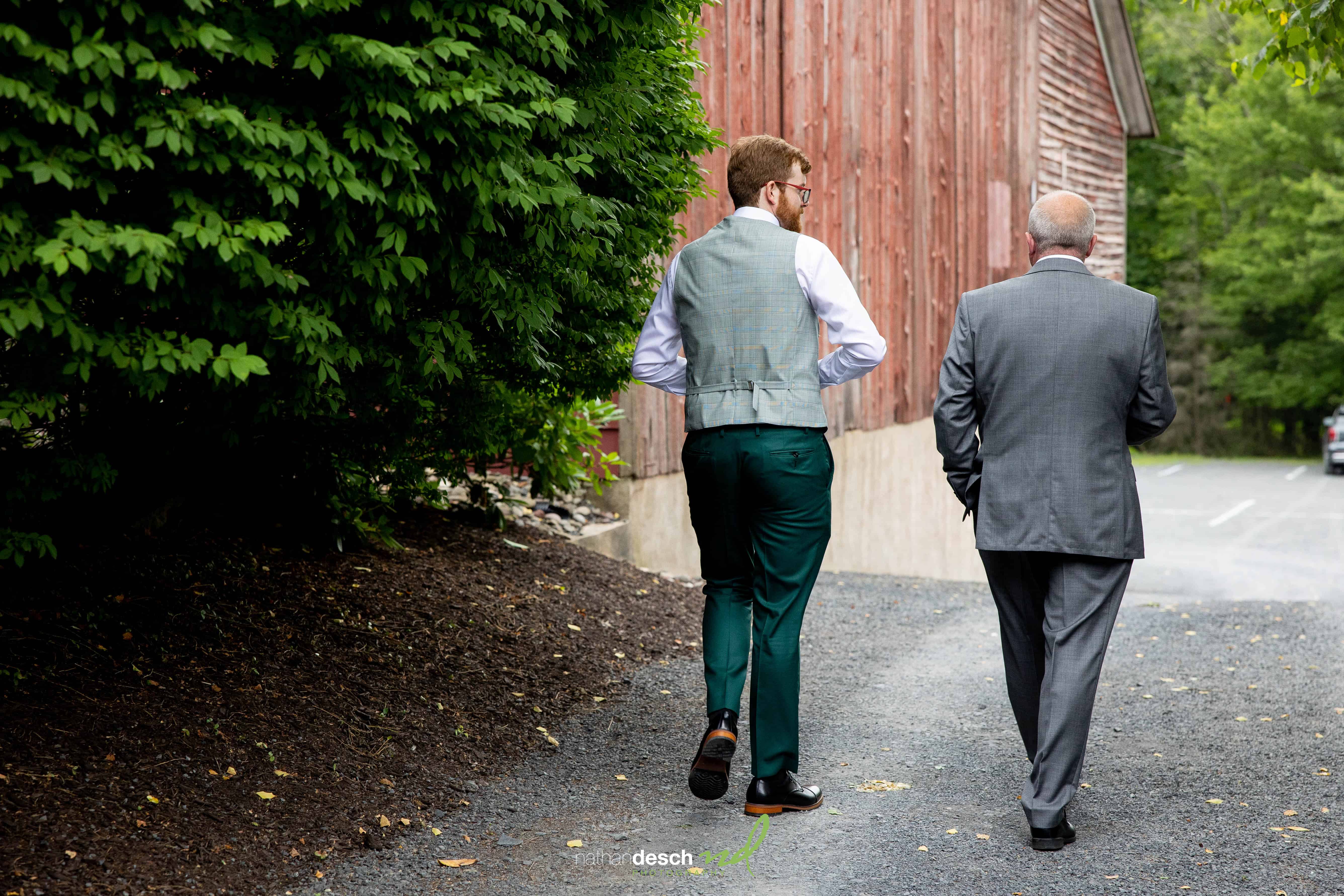 Two men in suits walk side by side on a gravel path next to a red barn and green trees at an outdoor wedding.