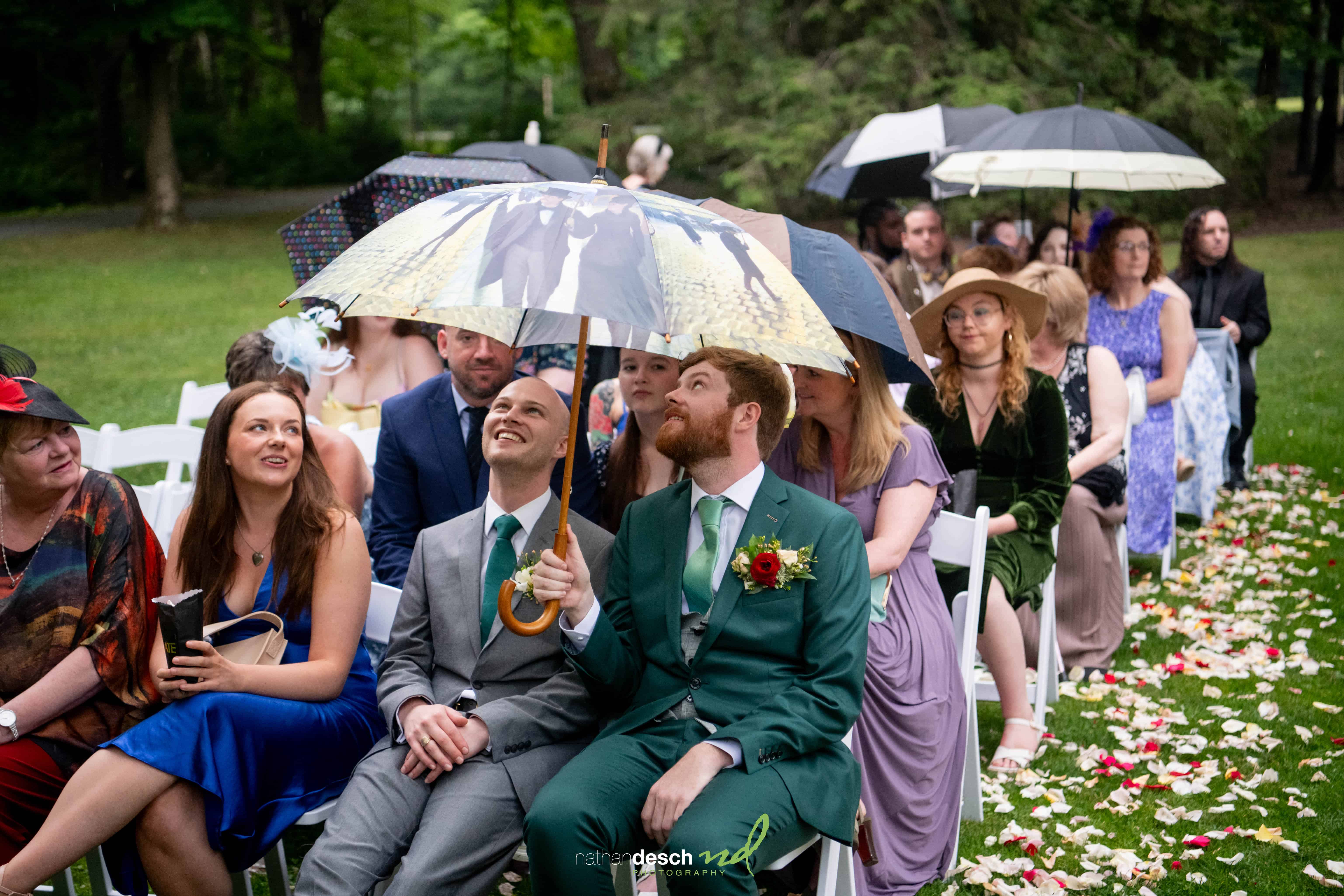 Wedding guests sit outdoors with umbrellas in the rain, smiling and chatting at Jake and Erika’s lush, green celebration.