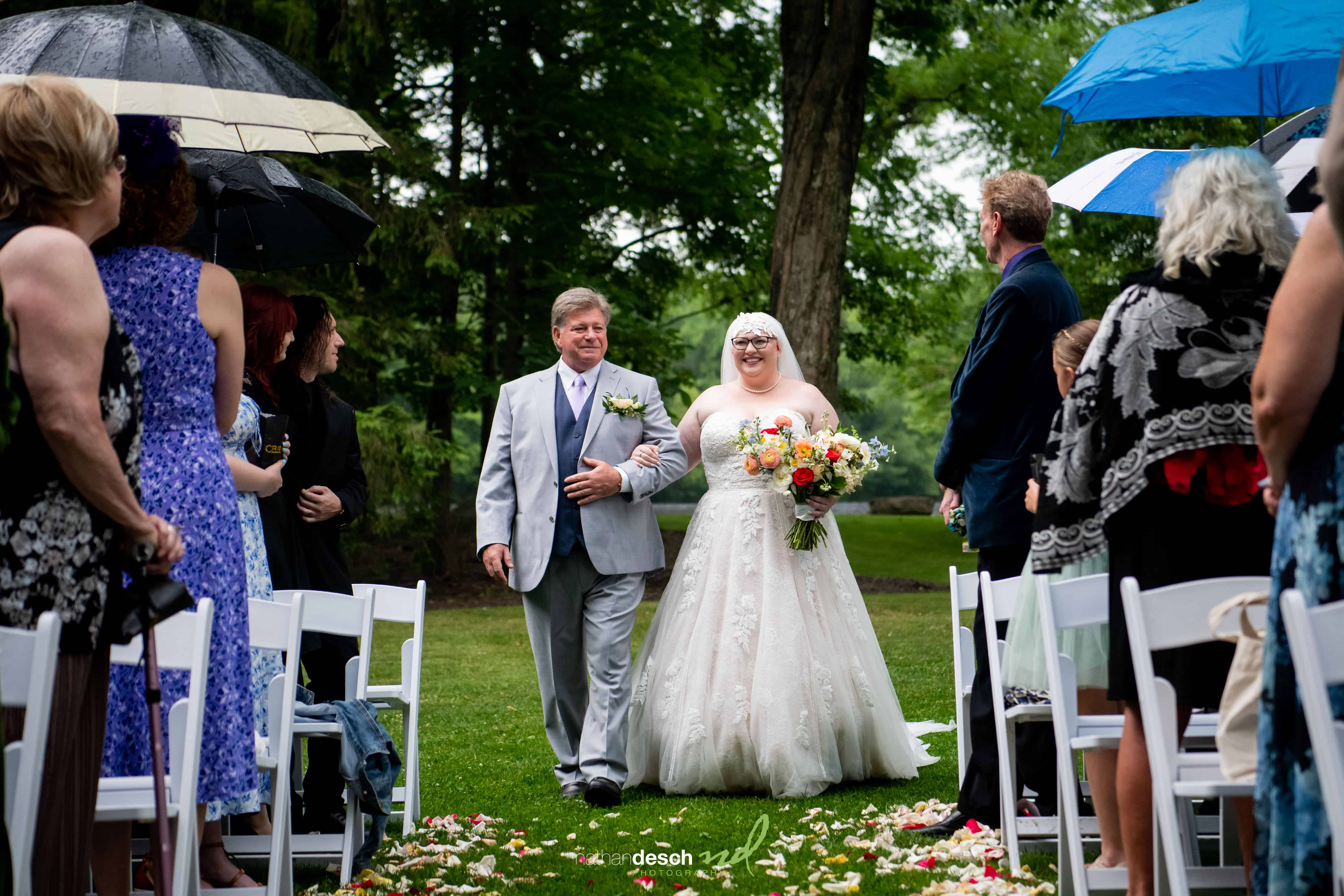 Bride in white gown walks down rainy outdoor aisle with father, smiling and holding colorful bouquet under umbrellas, wedding guests watching.