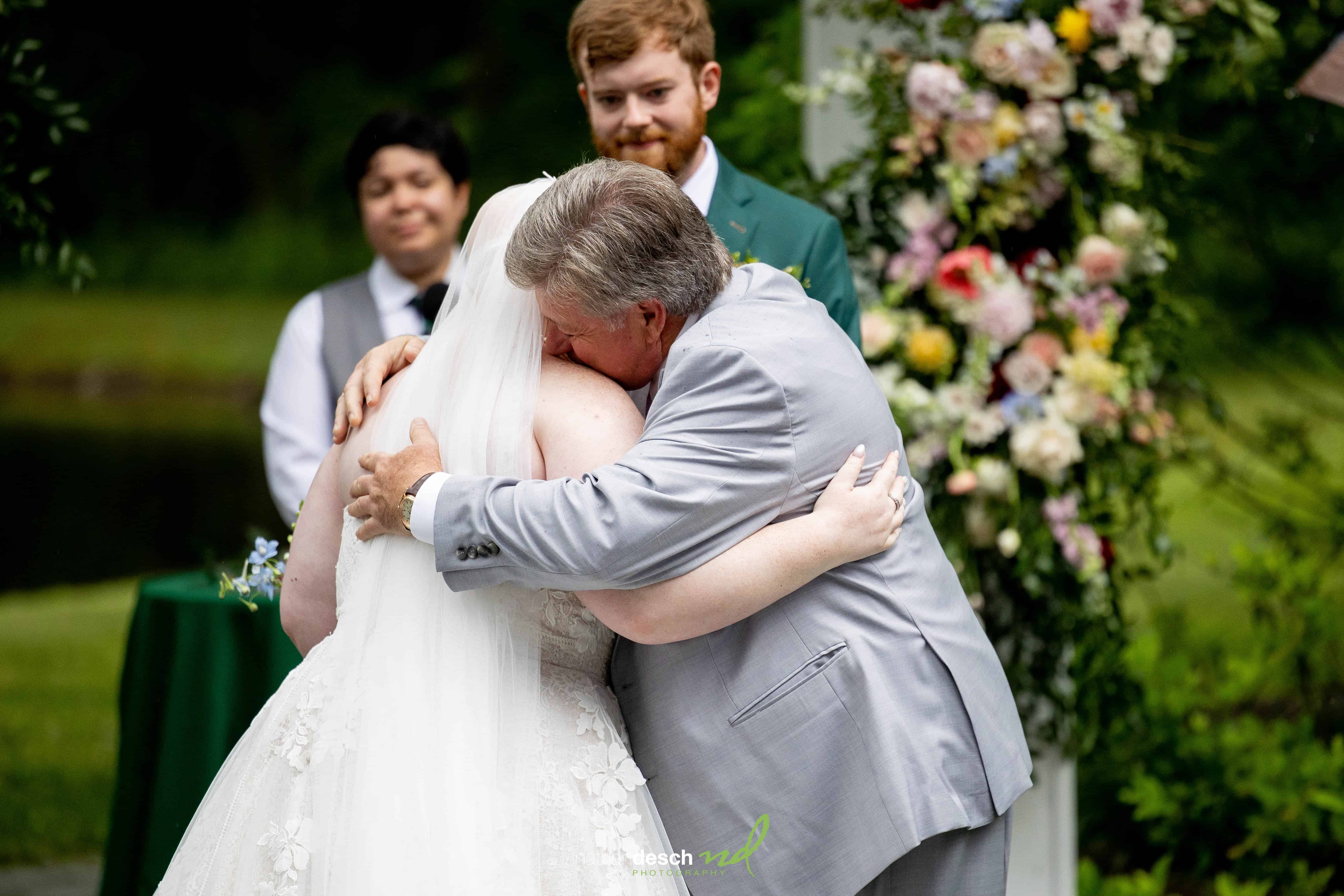 Bride in white dress hugs father in gray suit at outdoor wedding ceremony with floral decor and guests smiling nearby.