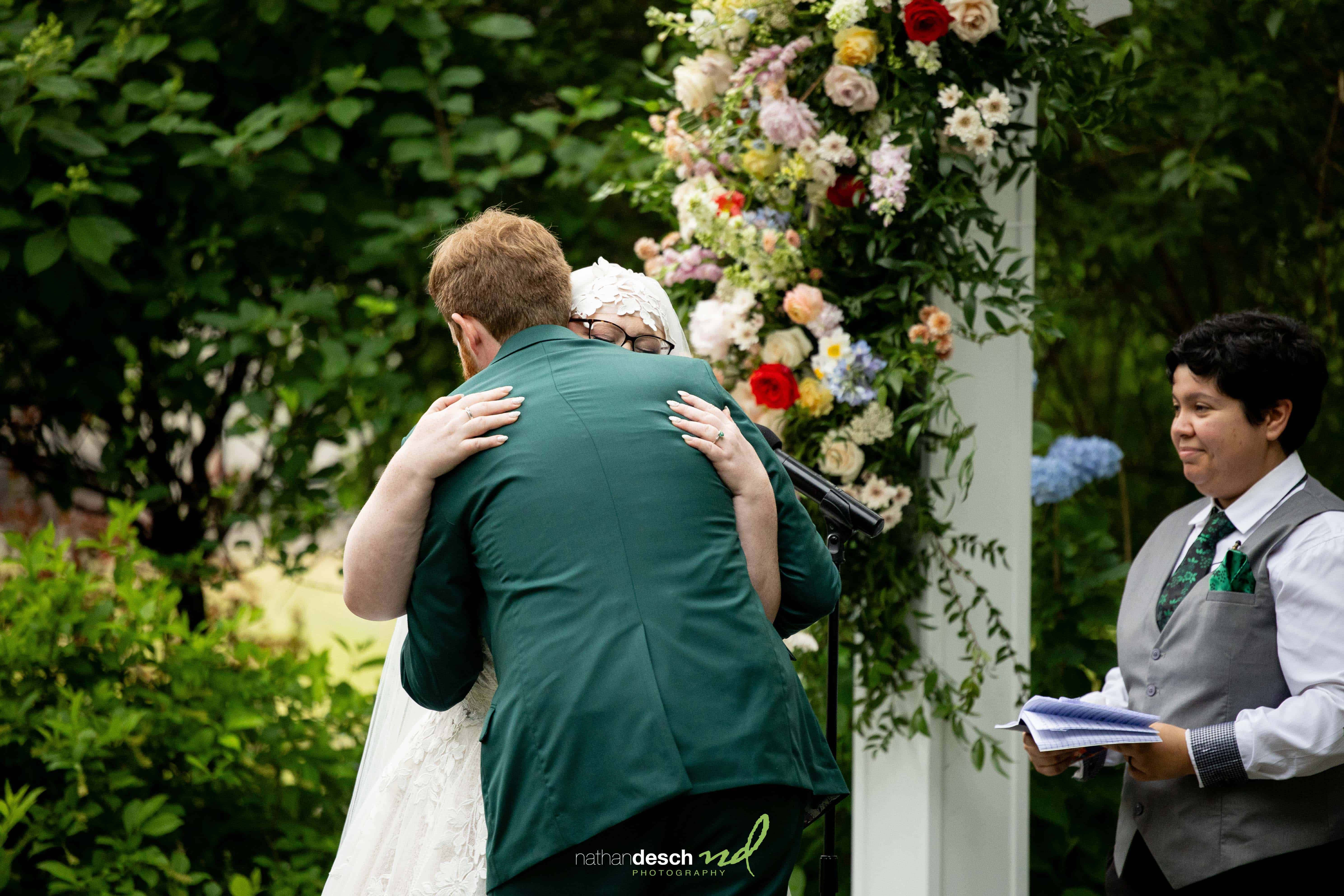 Bride and groom embrace beneath a floral arch during outdoor wedding ceremony with officiant, surrounded by lush greenery.