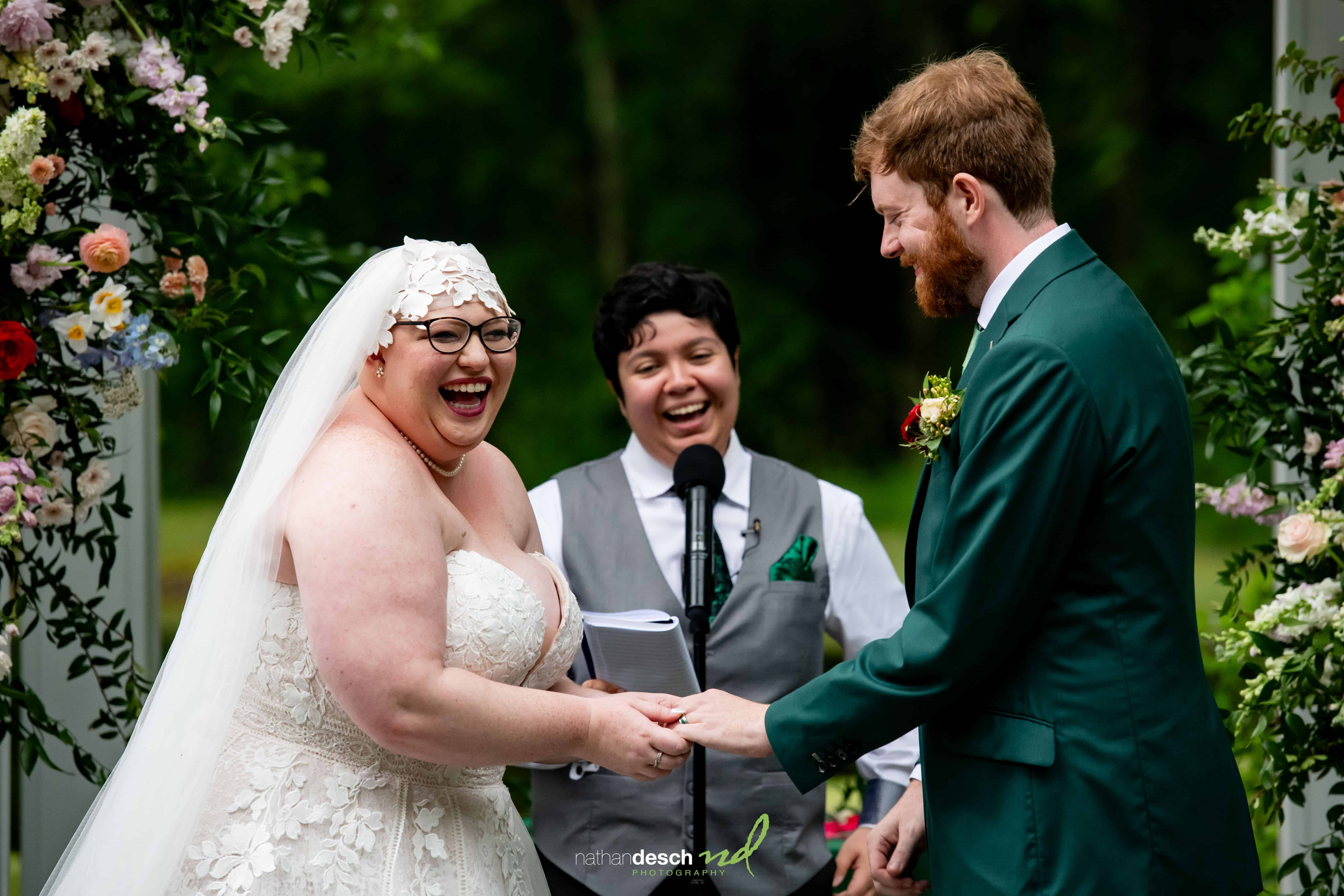 Bride and groom hold hands and smile at outdoor wedding ceremony, surrounded by colorful flowers and lush greenery.