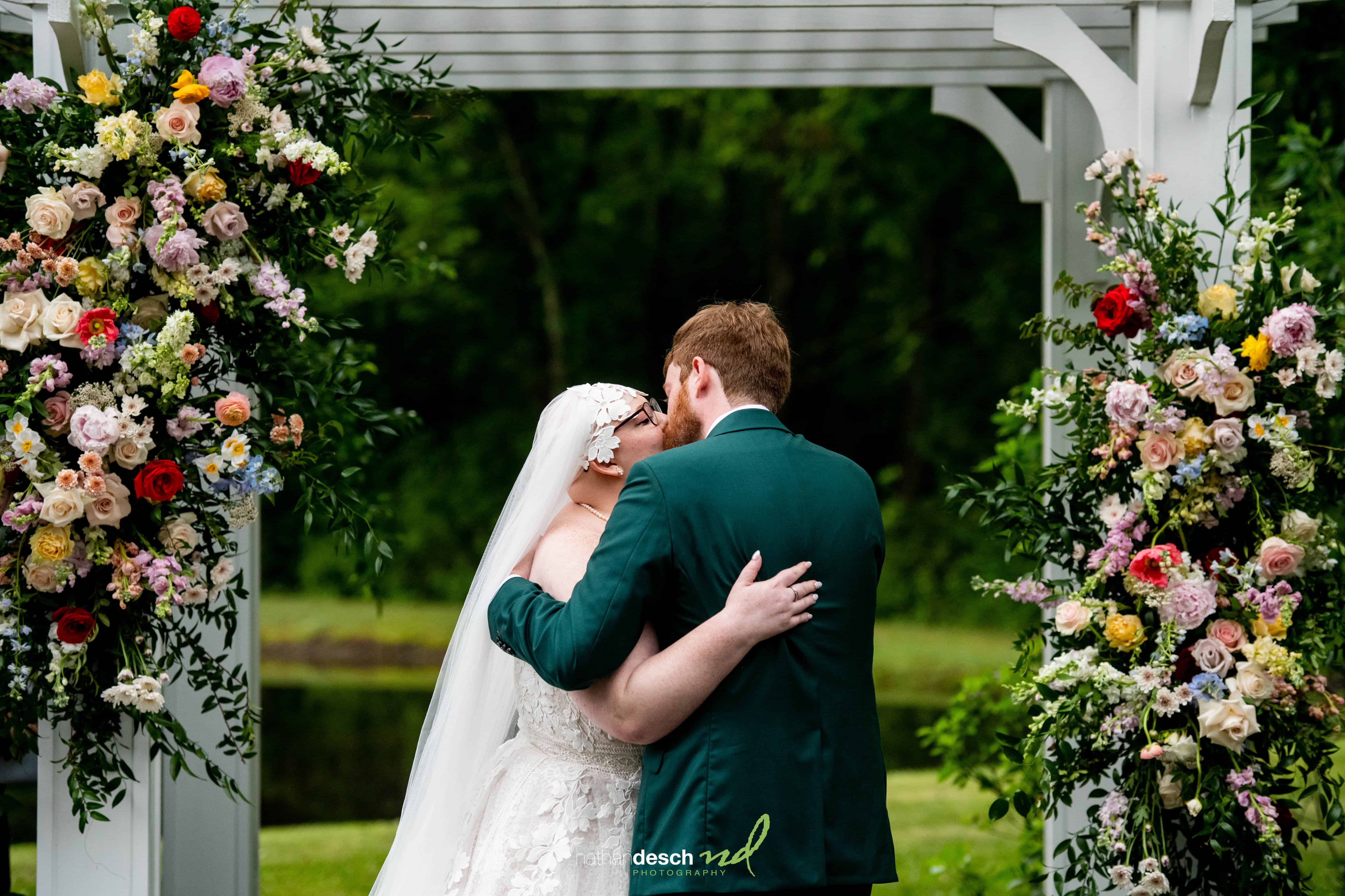 Bride and groom kiss under floral pergola, bride in white dress and veil, groom in dark green suit, lush greenery backdrop.