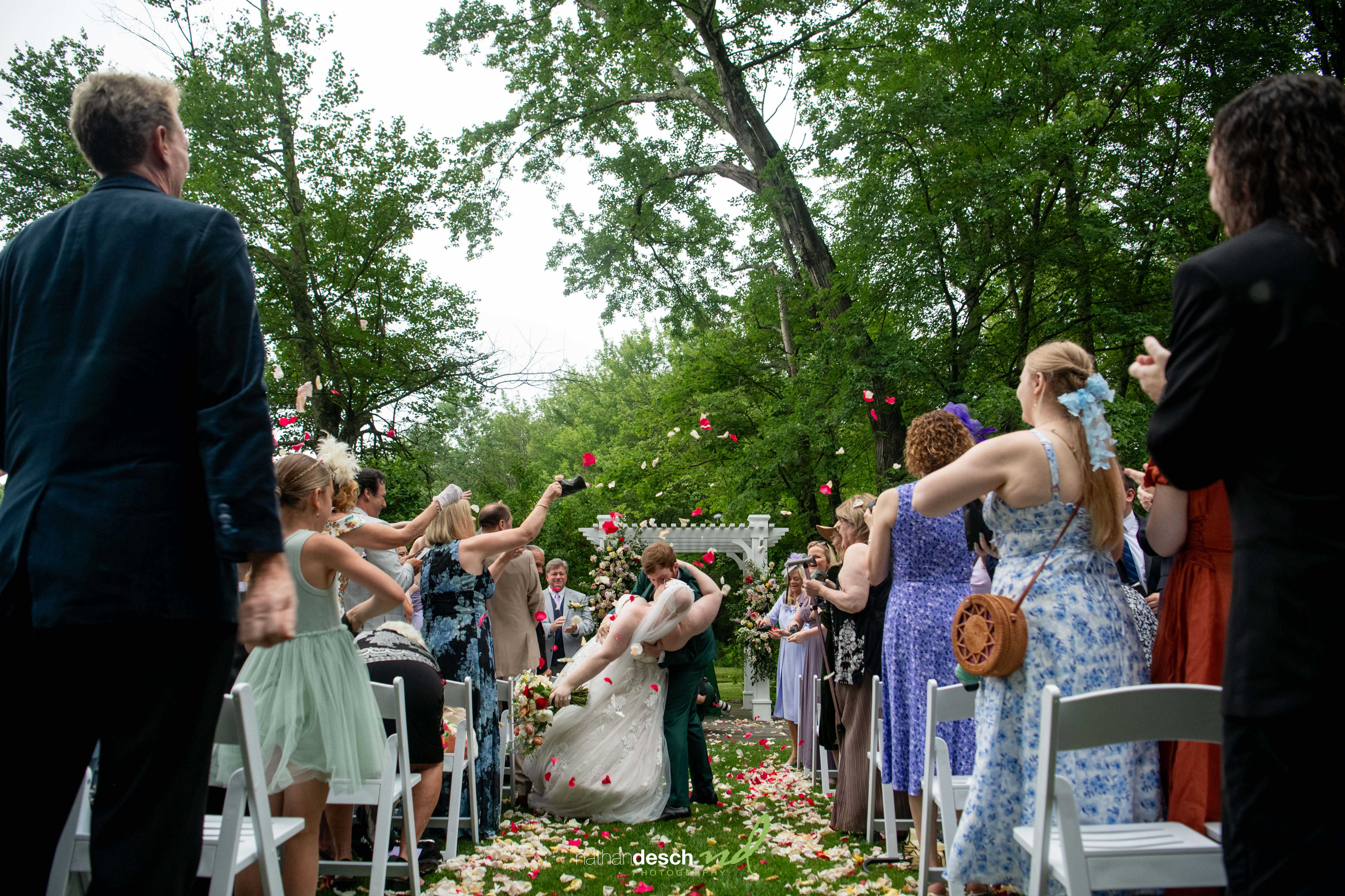 Bride and groom share a kiss under trees as wedding guests cheer and toss colorful flower petals down the aisle.