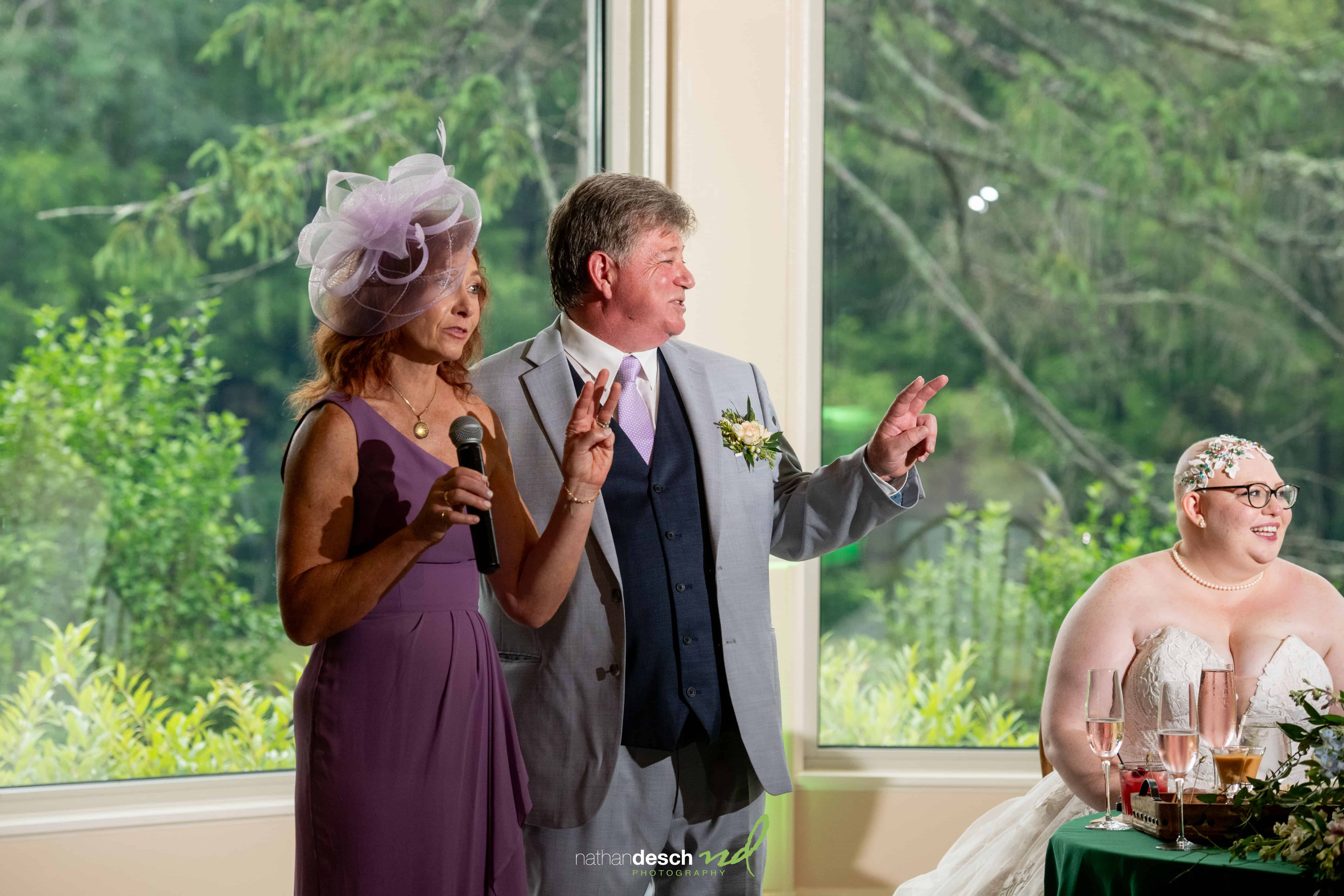 Wedding guests in vibrant outfits laughing with the bride at a sunlit table with lush greenery outside large windows.