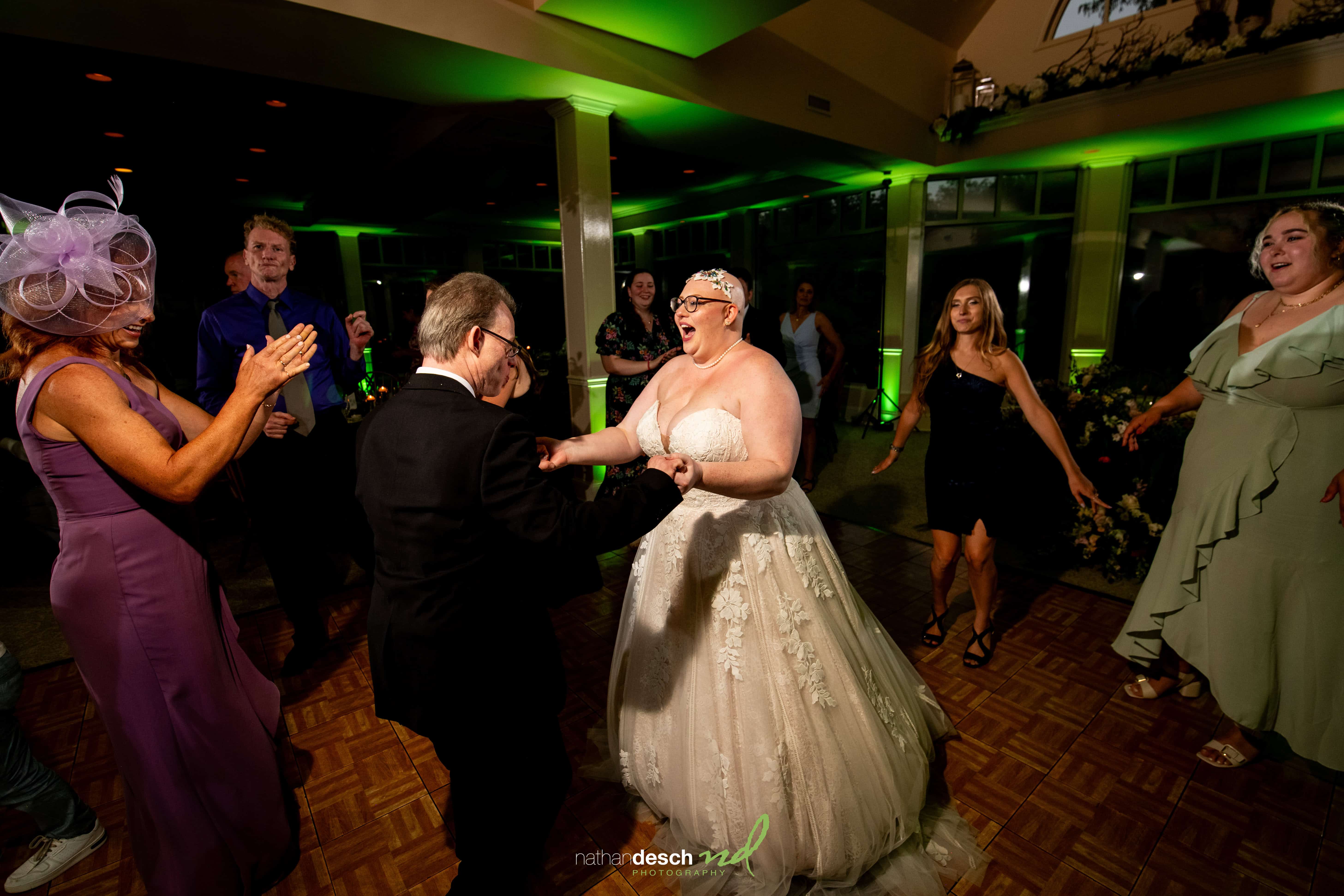 Jake and Erika share a joyful dance on a wooden floor as wedding guests in formal attire smile under green reception lights.