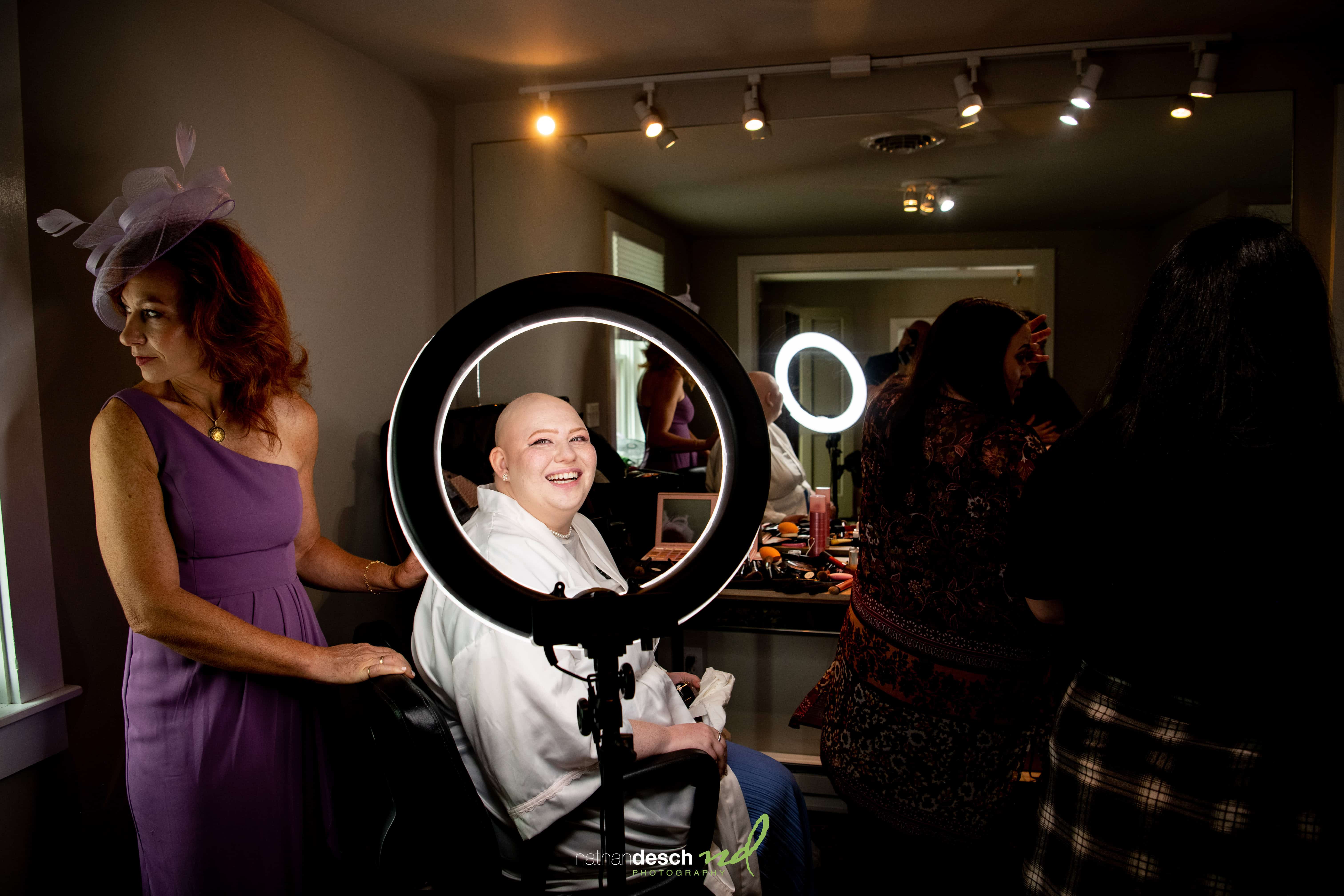 Bald woman smiling under ring light as wedding party prepares, with Jake, Erika, and a guest in a purple dress nearby.
