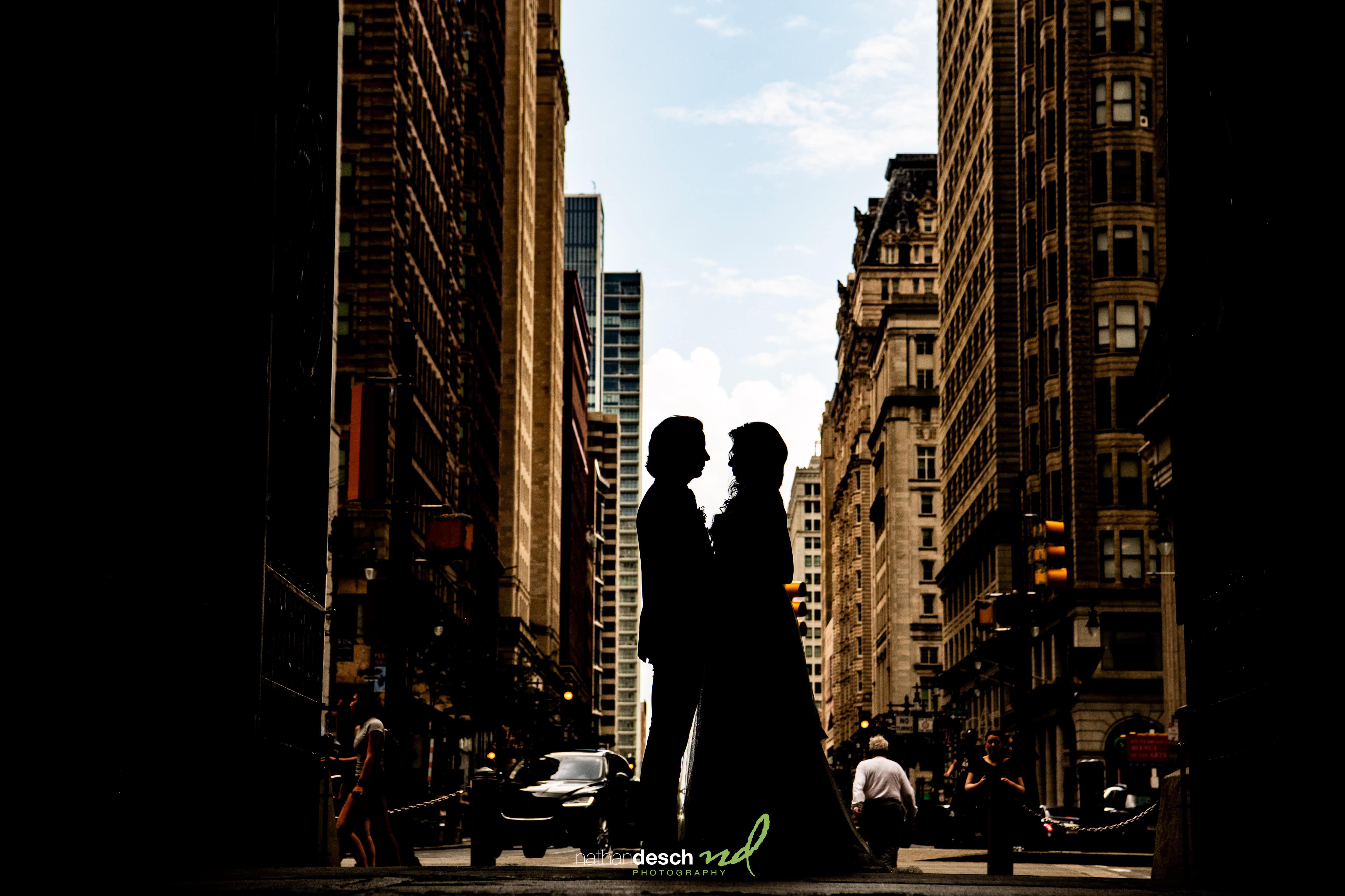 Silhouette of bride and groom facing each other on a city street with tall buildings, cars, and pedestrians at sunset.