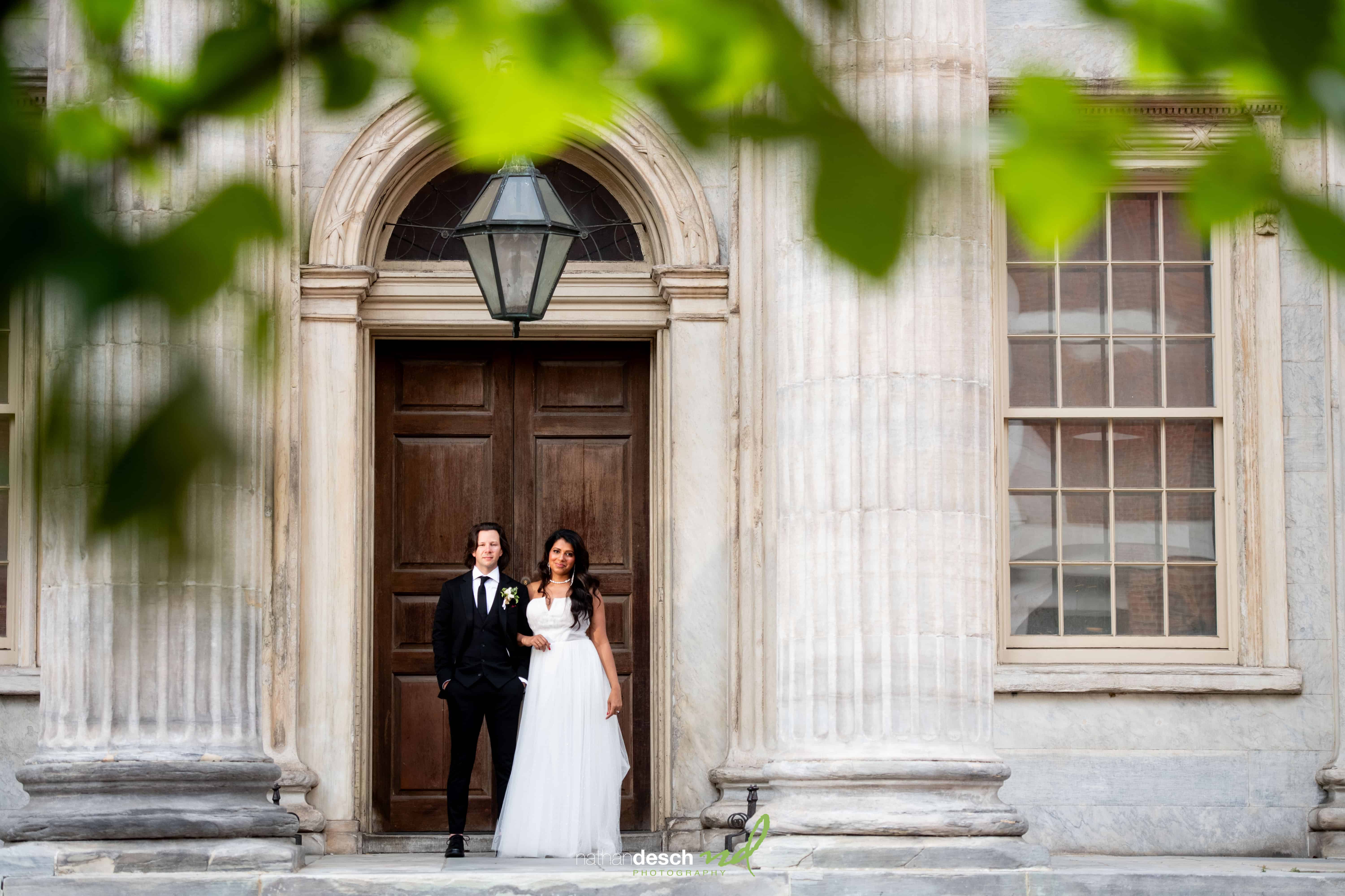Bride and groom posing together by grand wooden doors and stone columns, surrounded by lush greenery on their wedding day.
