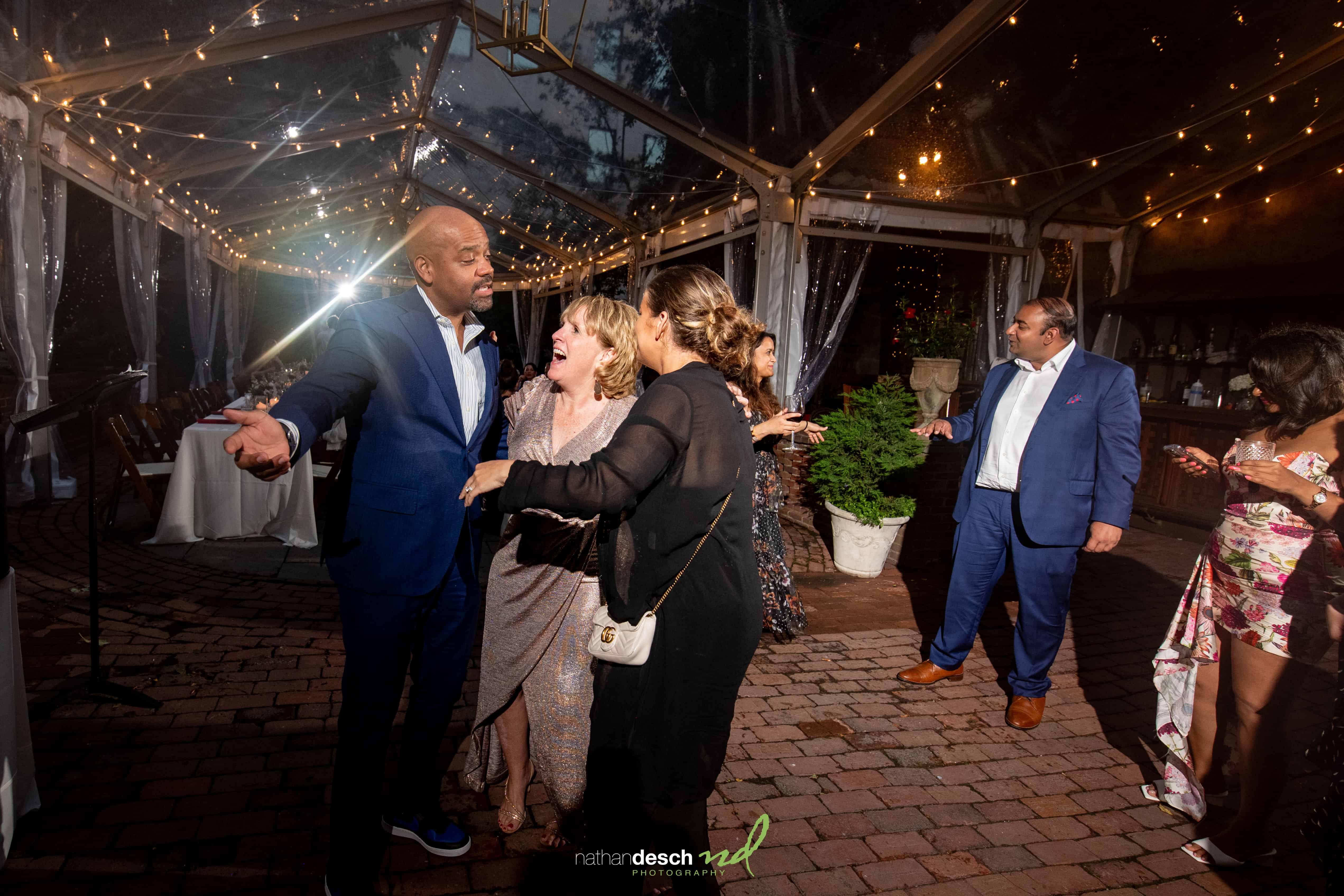 Brian and Anusha dancing and laughing with guests in formal wear under string lights at an outdoor wedding reception at night.