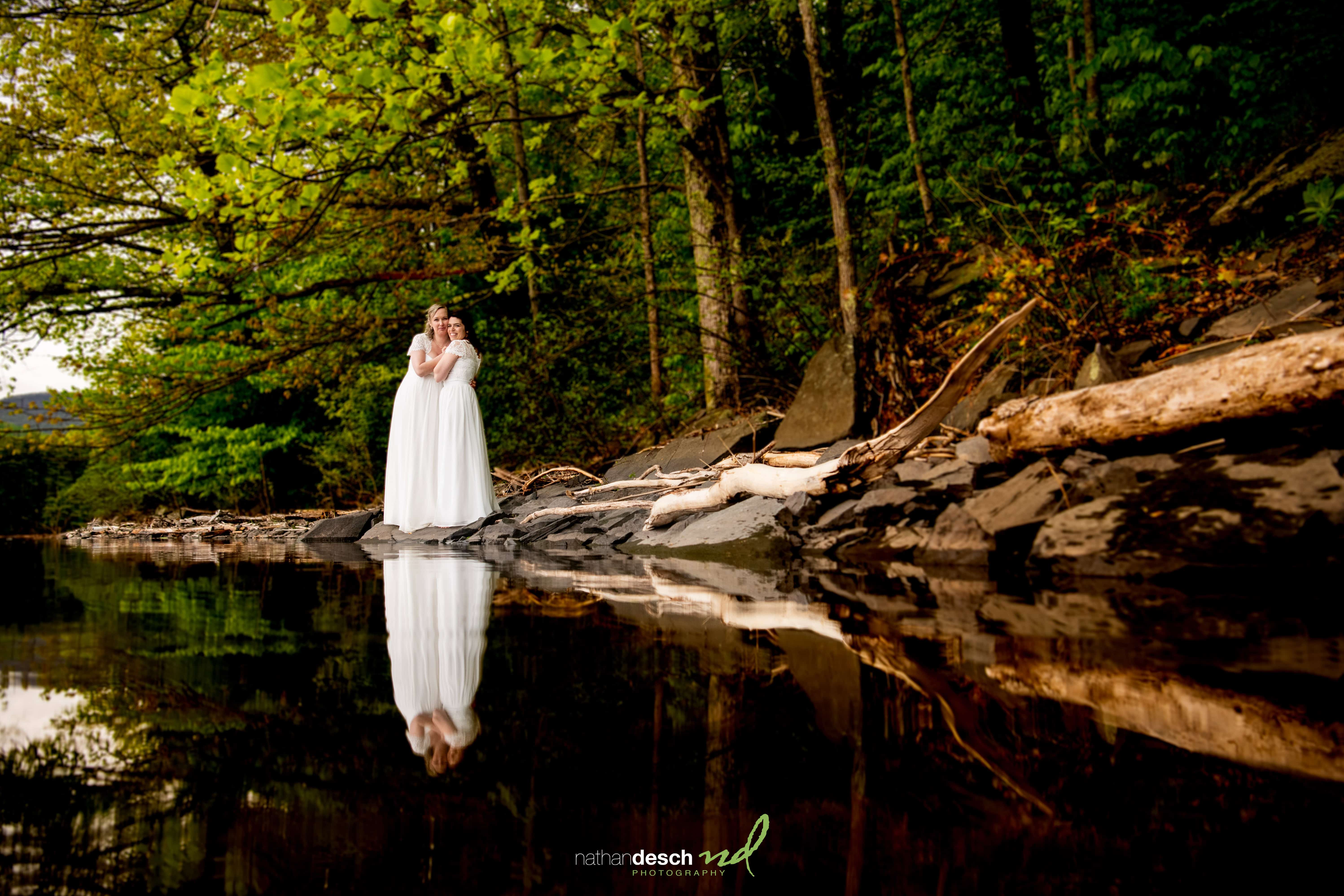 Two brides in white dresses embrace by a calm riverbank in the Catskills, surrounded by lush green trees and reflections.