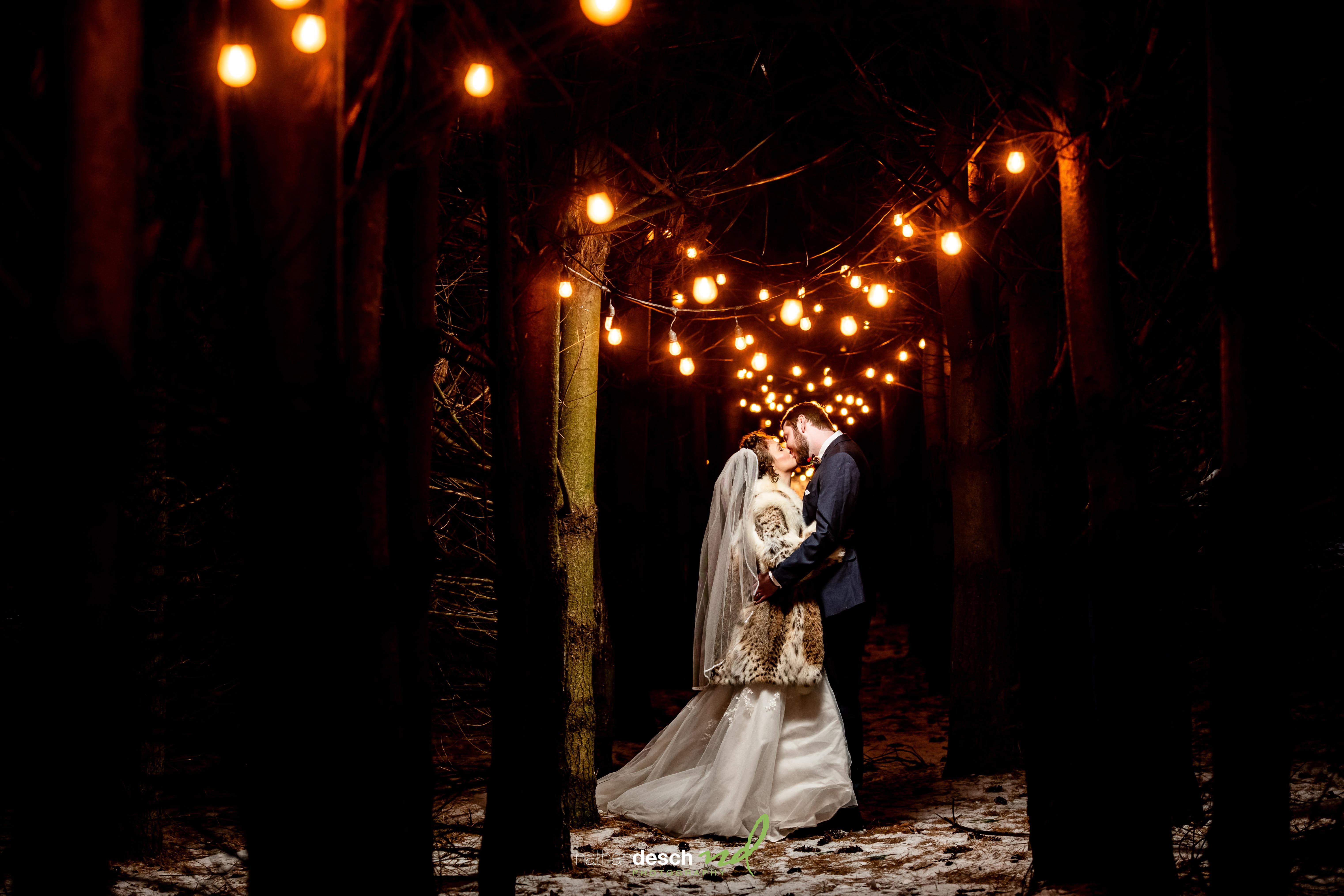 Bride and groom embrace on a snowy forest path at night under glowing string lights, creating romantic winter wedding inspiration.