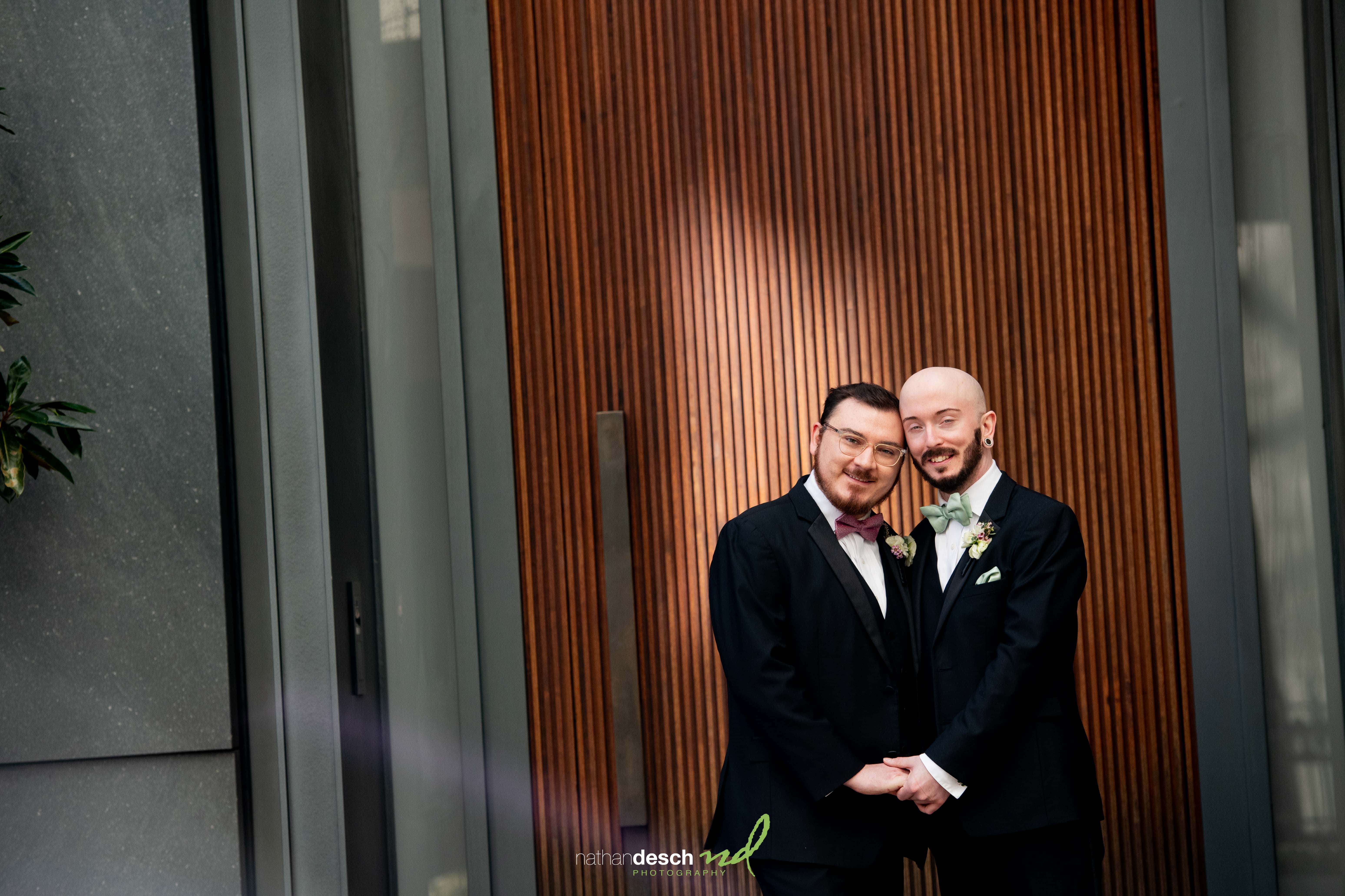 Two grooms smiling and holding hands in suits with boutonnières, celebrating their spring wedding at Tendenza venue.