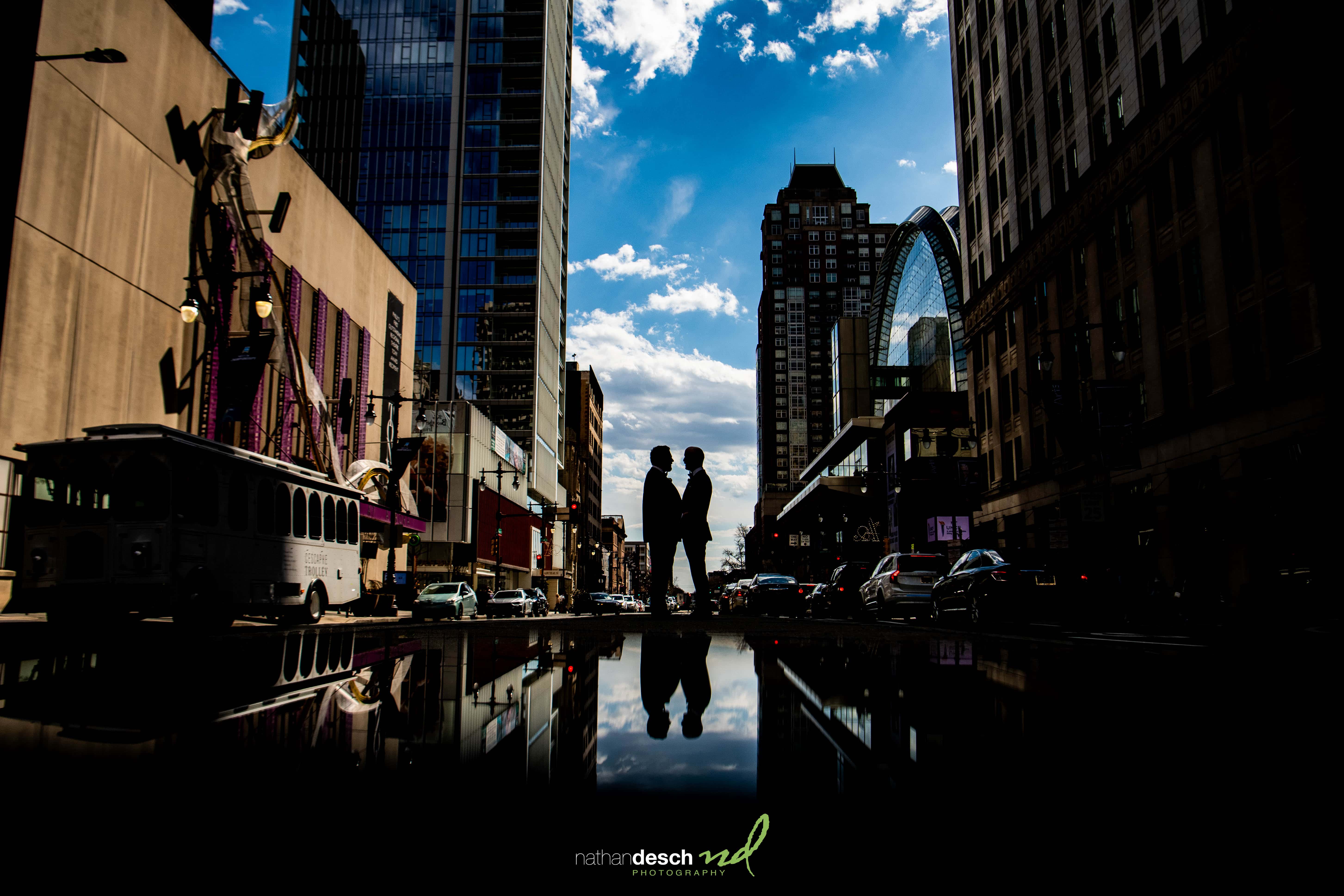Silhouetted wedding couple embracing on a city street at Tendenza, reflected on wet pavement with cloudy blue sky overhead.