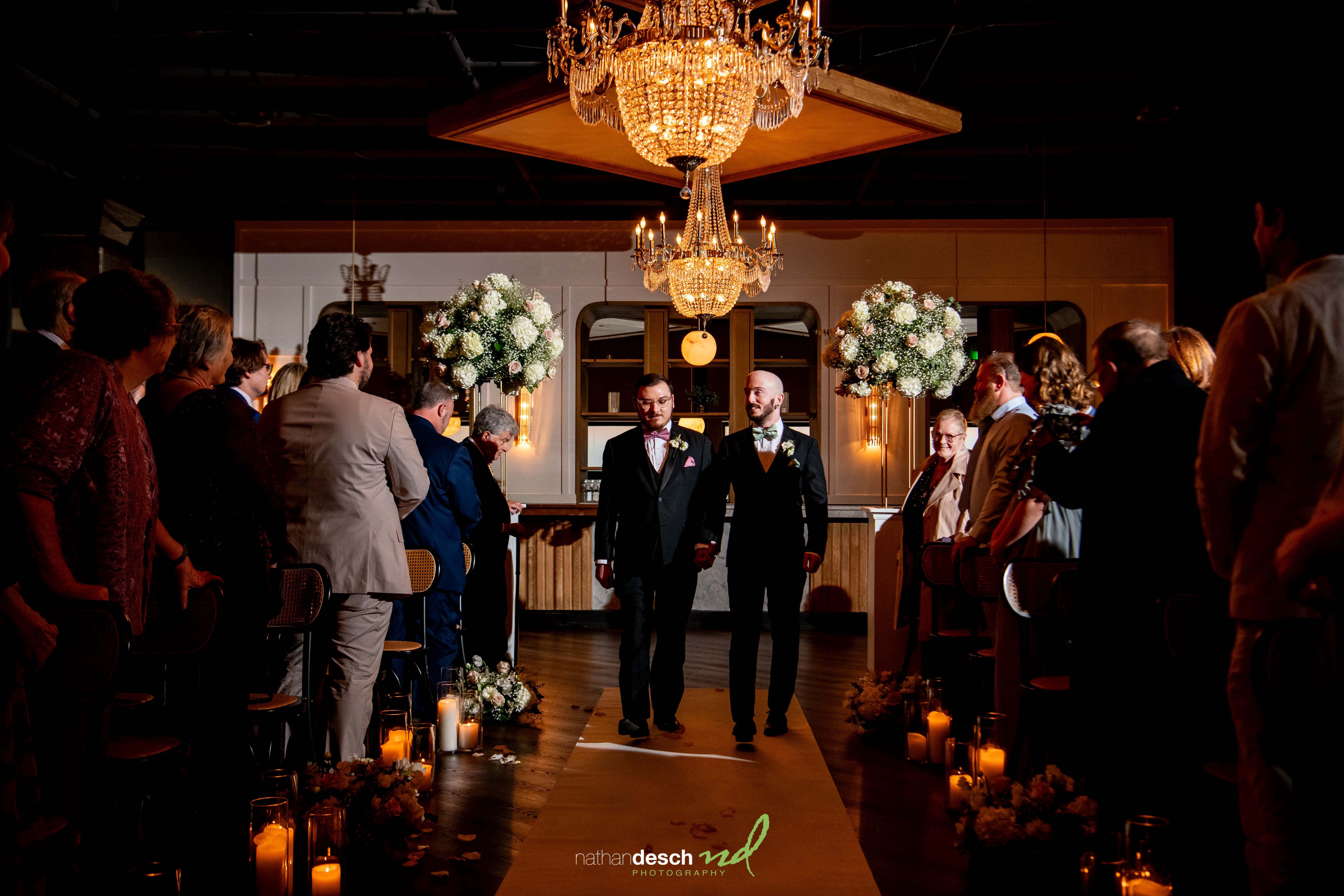 Two grooms in tuxedos walk hand-in-hand down a candlelit aisle with flowers at Tendenza spring wedding ceremony.