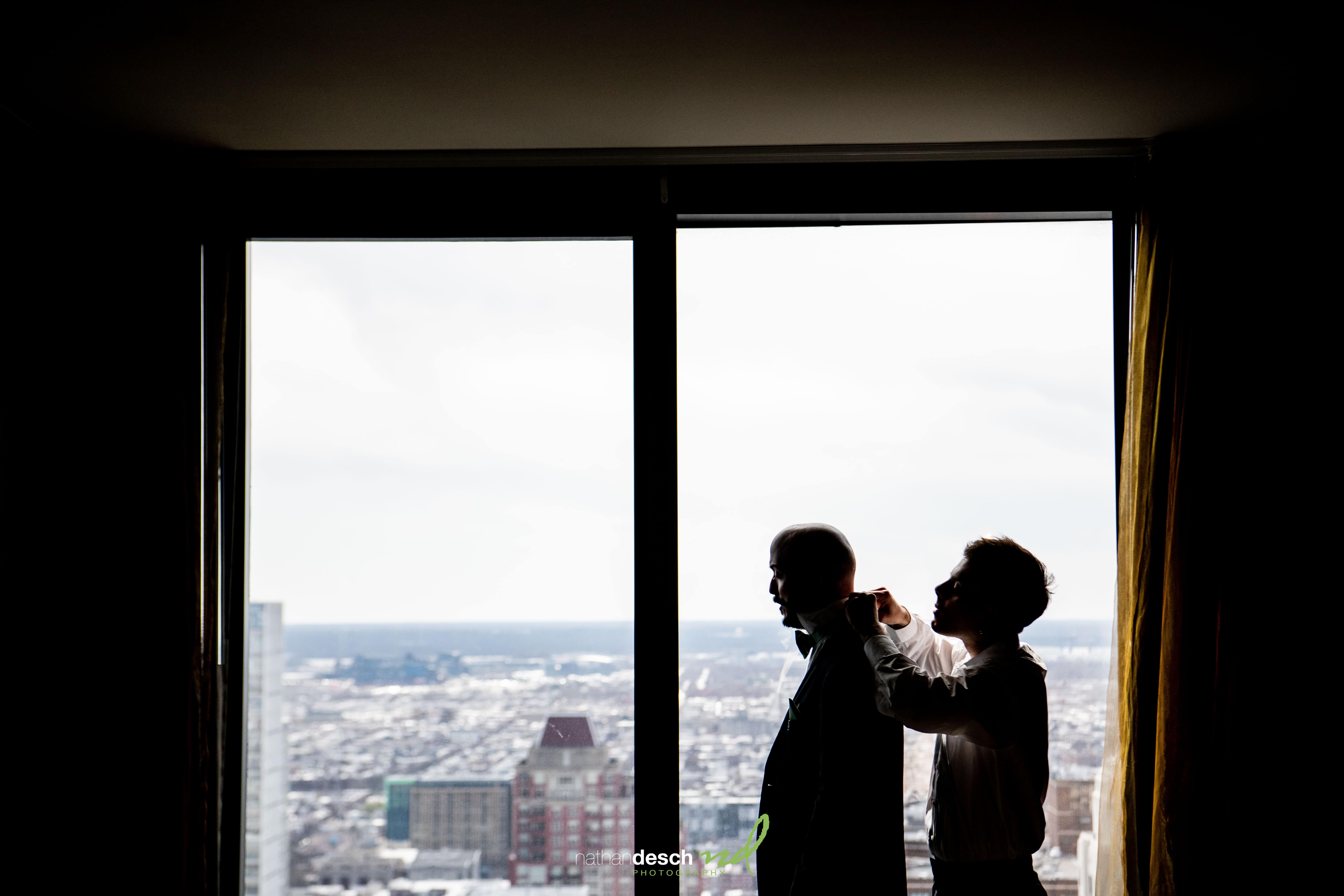 Groom getting bow tie adjusted by best man near large window with city view at spring wedding at Tendenza Philadelphia.