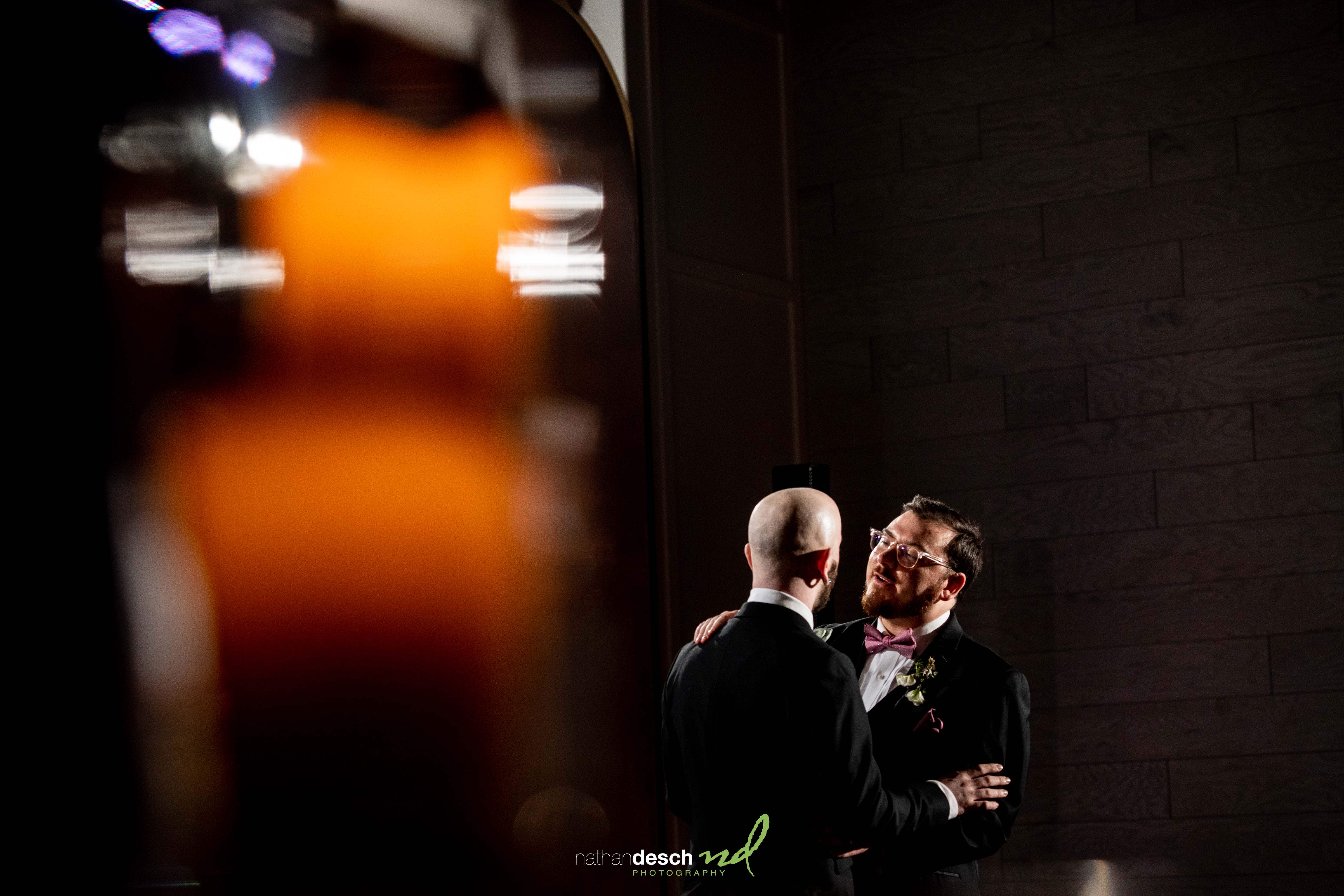 Grooms share a romantic first dance at Tendenza wedding, one in glasses and pink bow tie, soft spring lighting, blurred foreground.
