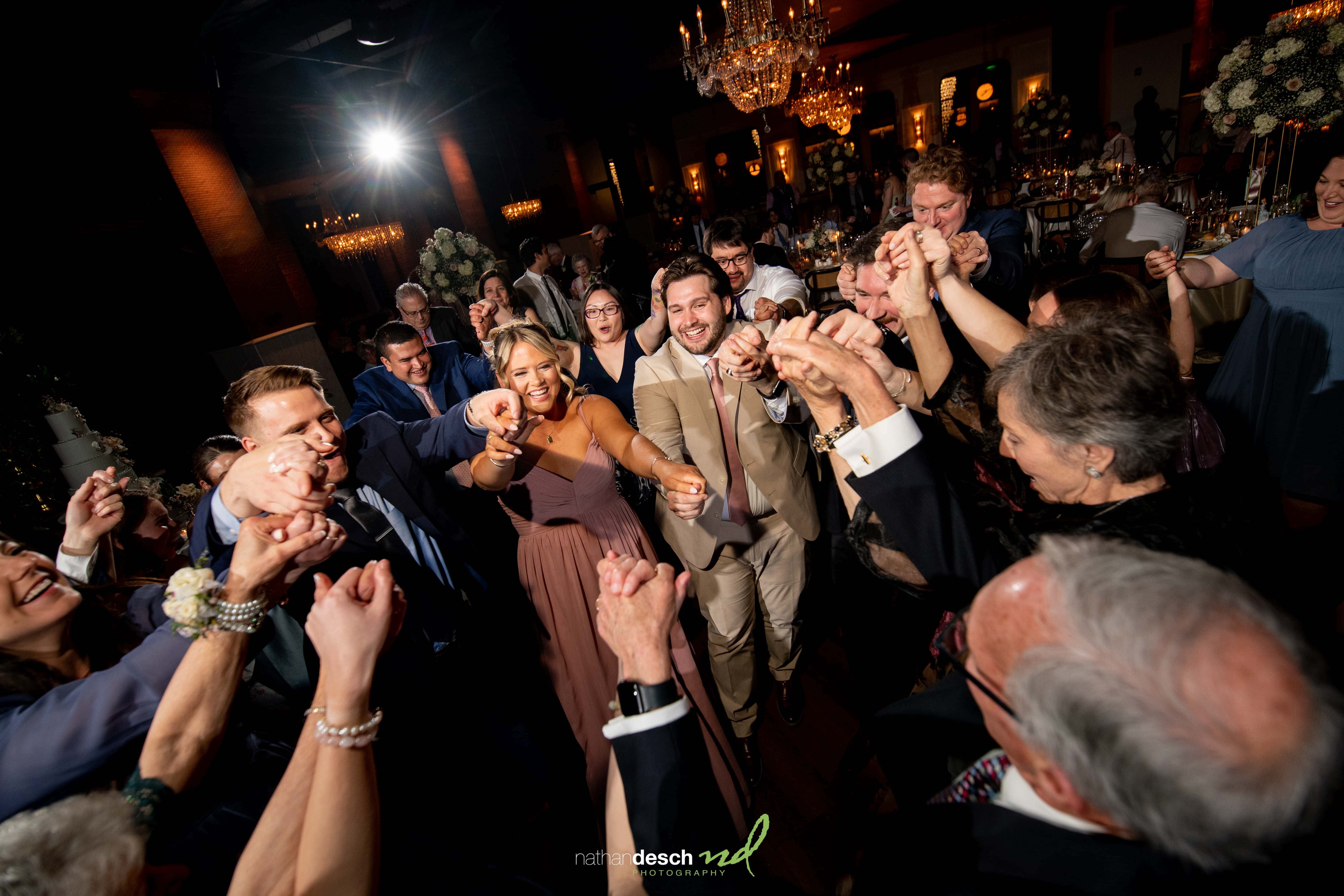 Guests in formal wear dance in a circle, smiling and holding hands at Tendenza spring wedding reception under chandeliers.