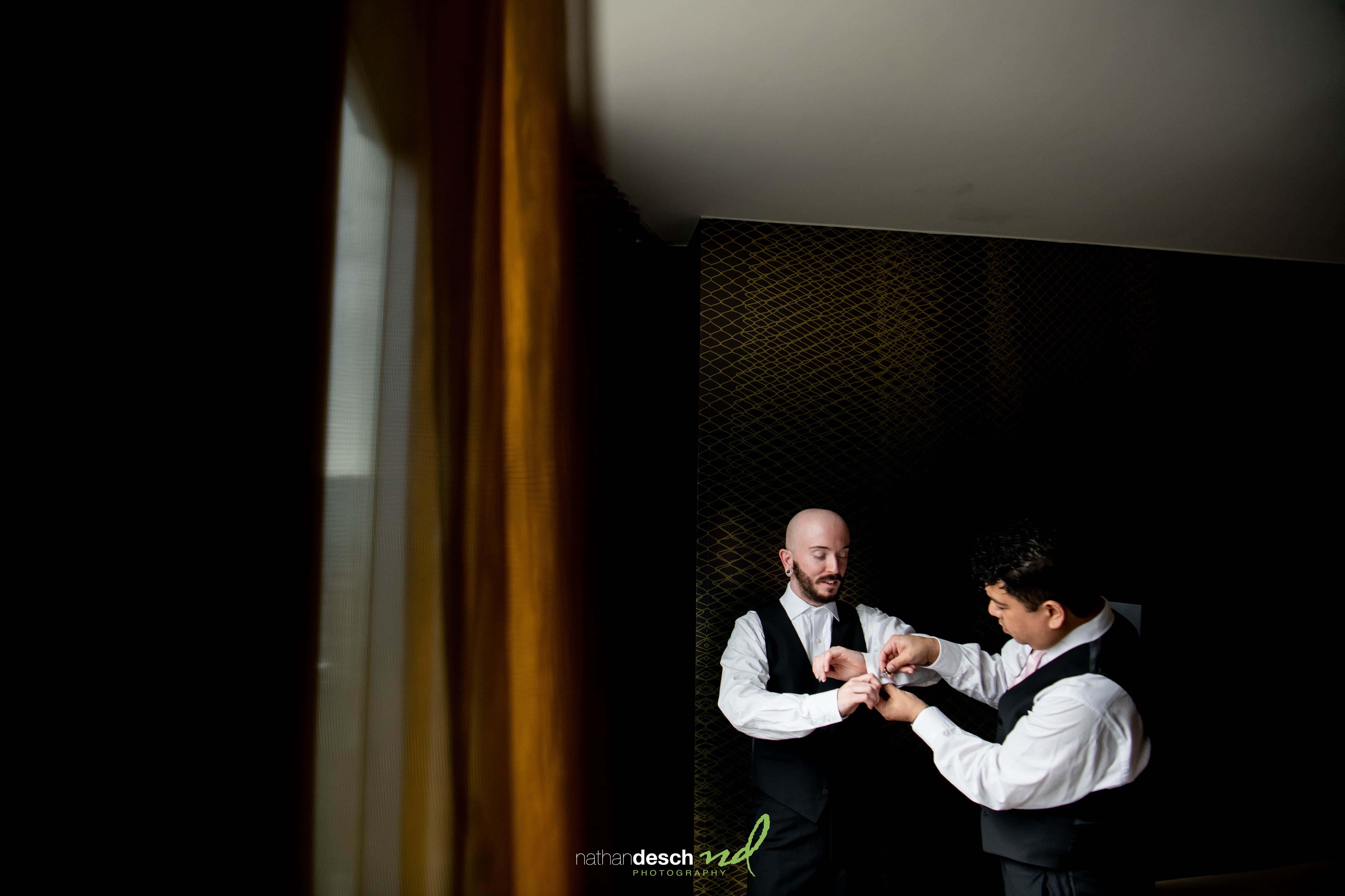 Groom and best man in formal attire, smiling as they adjust cufflinks by a window at Tendenza wedding venue.