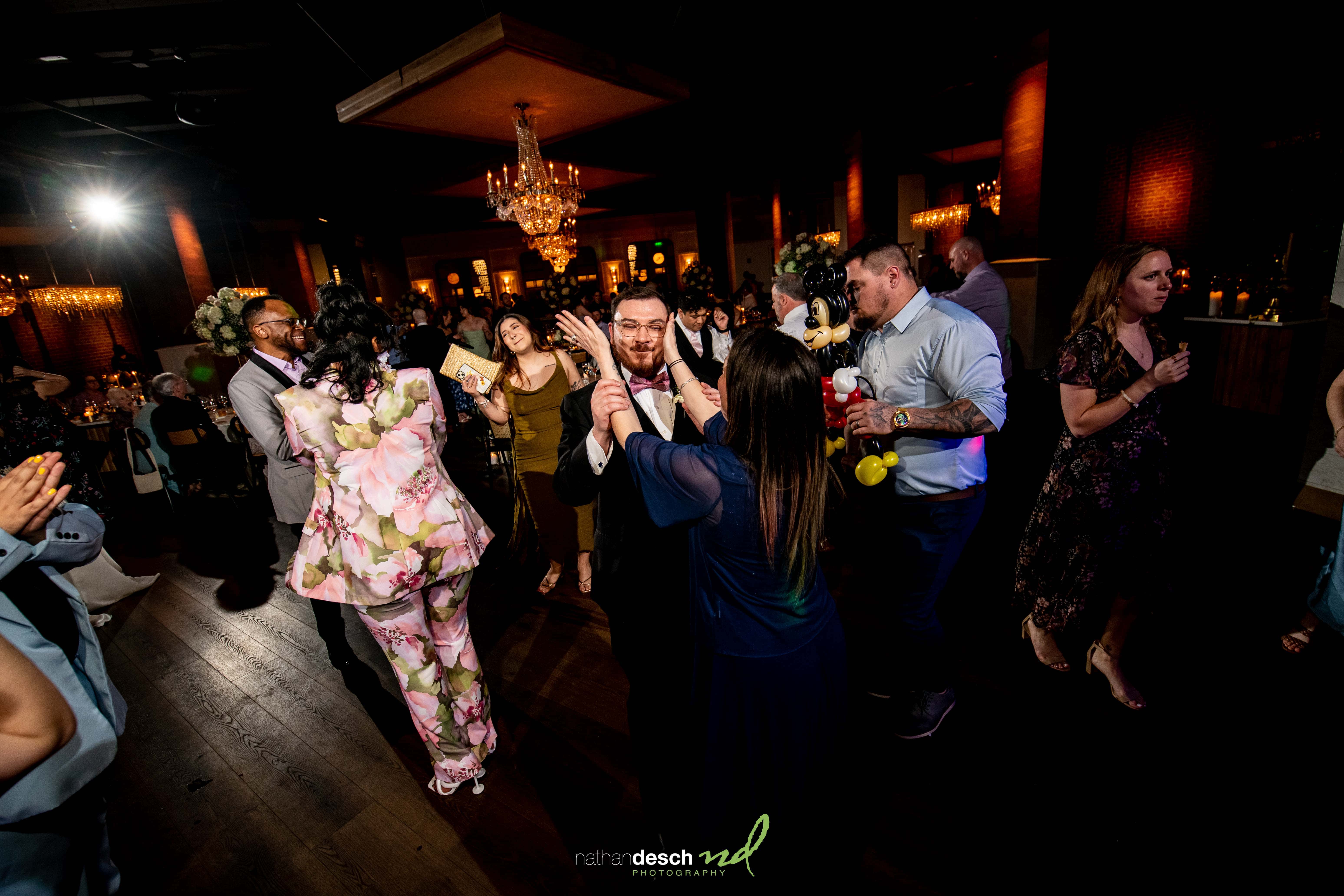 Guests in elegant attire dance joyfully under chandeliers at a vibrant spring wedding reception at Tendenza ballroom.