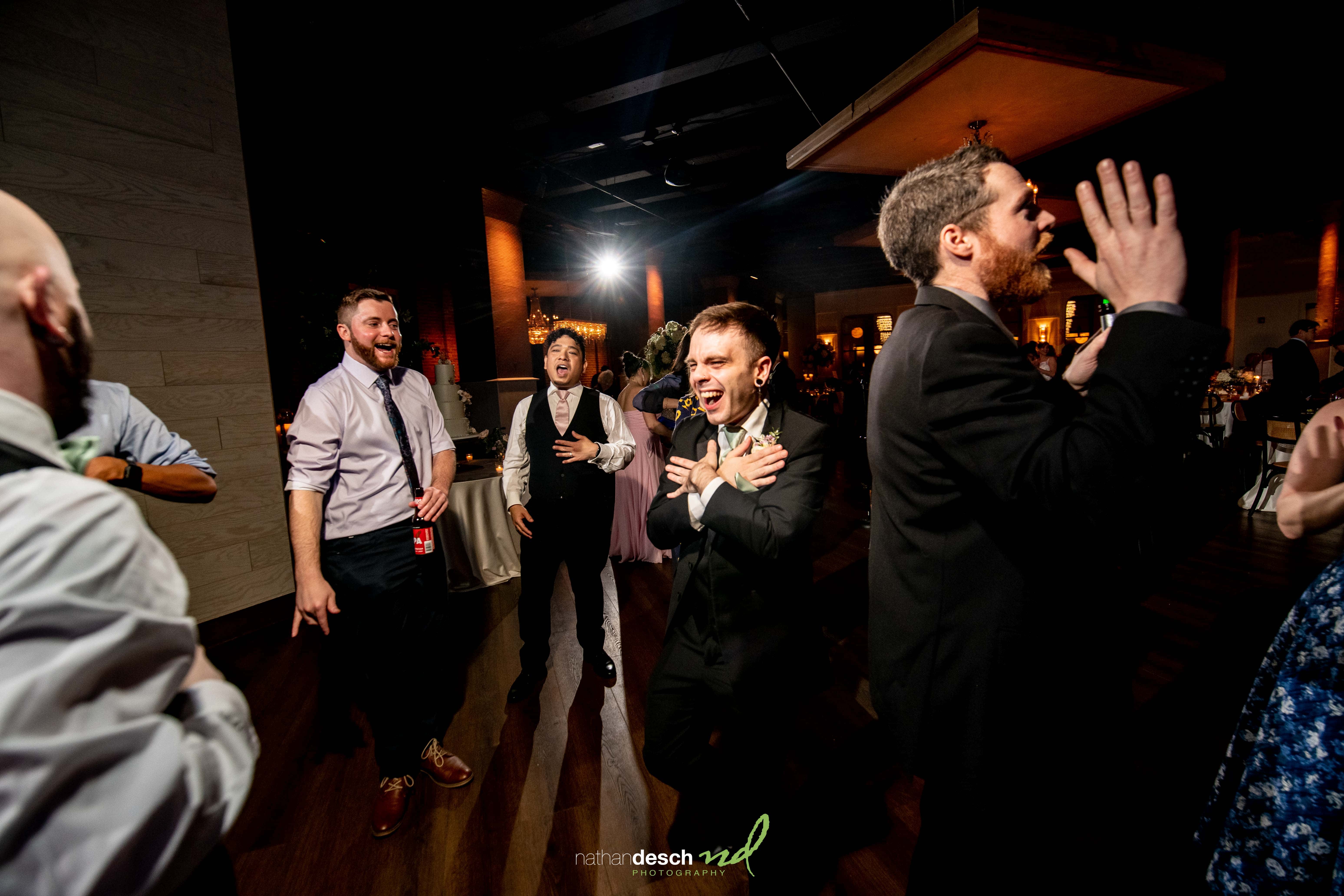 Groomsmen in suits laughing and dancing on a dimly lit dance floor during a lively spring wedding reception at Tendenza.