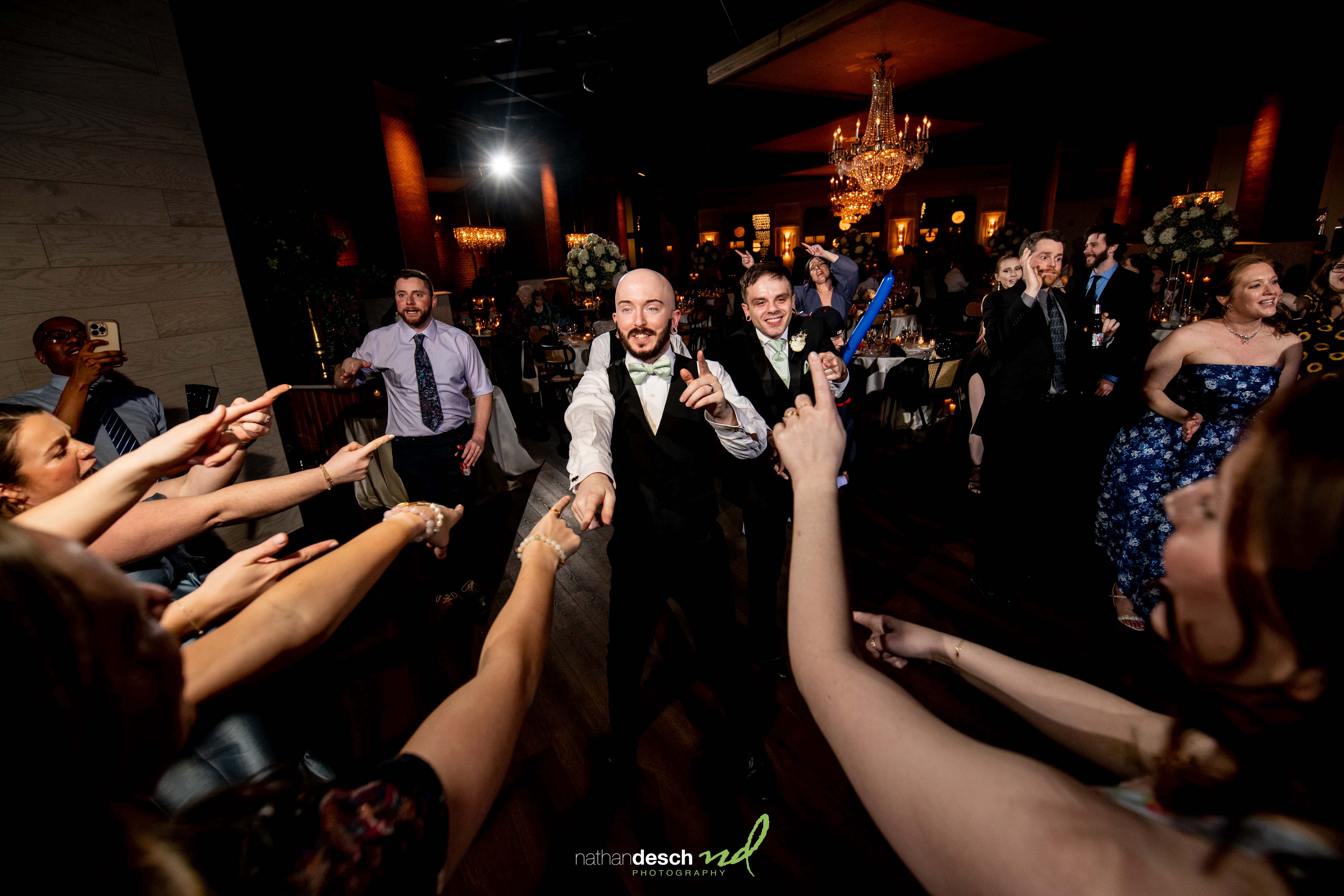 Two grooms in tuxedos dancing joyfully with guests at a spring Tendenza wedding, under glowing chandeliers and festive lights.