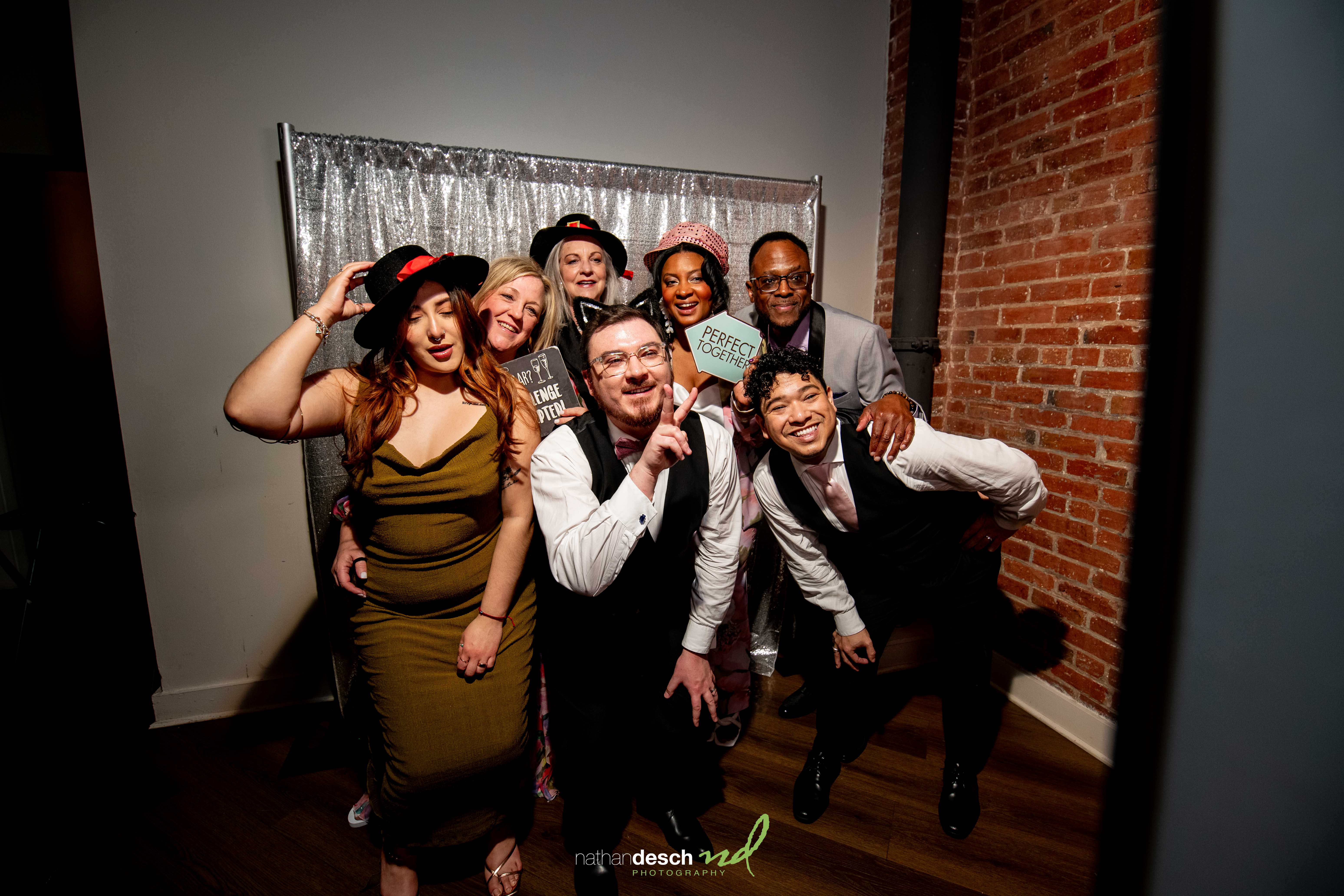 Group of guests in semi-formal outfits pose with props, smiling by silver sequin backdrop at Tendenza spring wedding.
