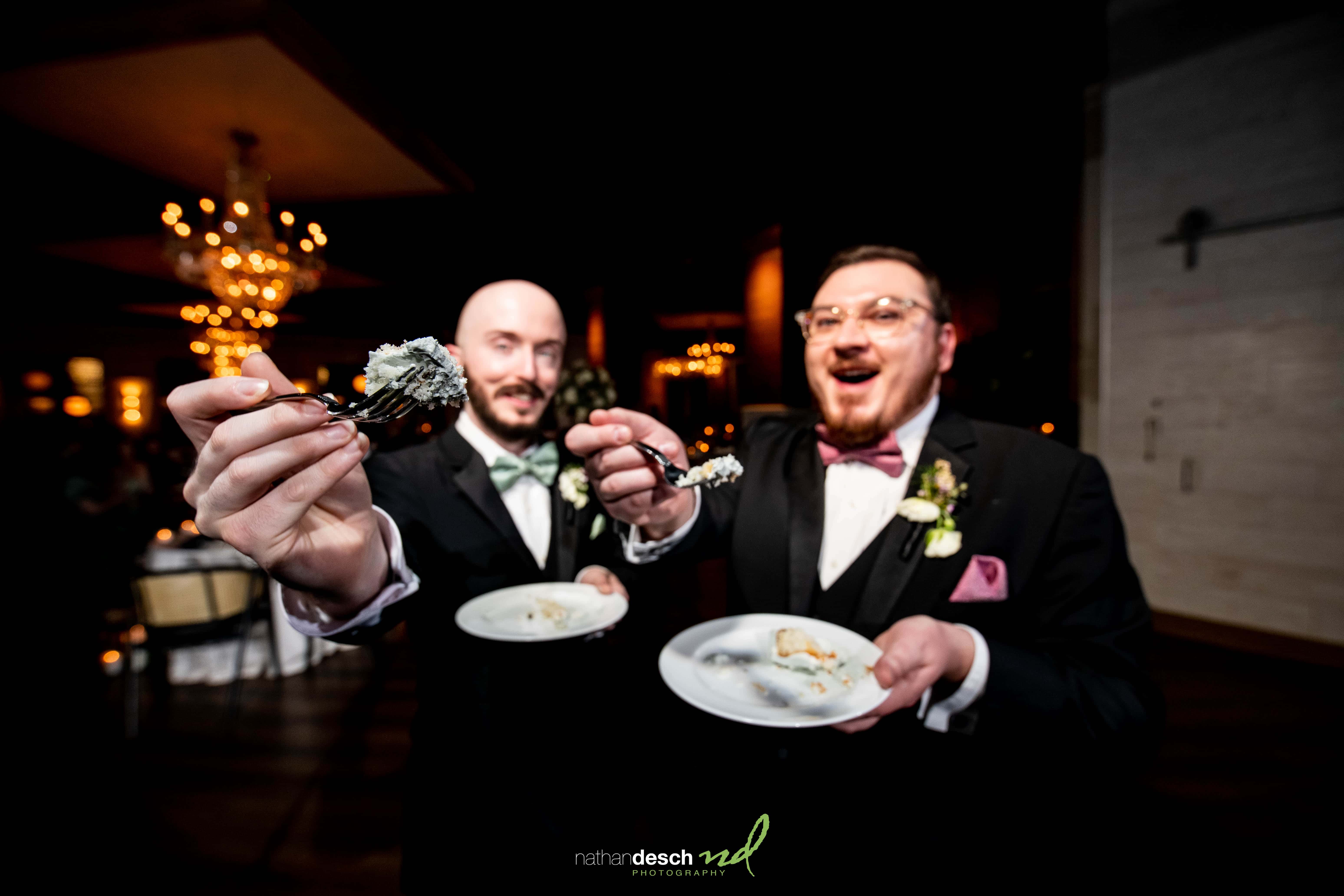Two grooms in tuxedos share wedding cake at Tendenza, smiling and celebrating under chandeliers in a festive, elegant venue.