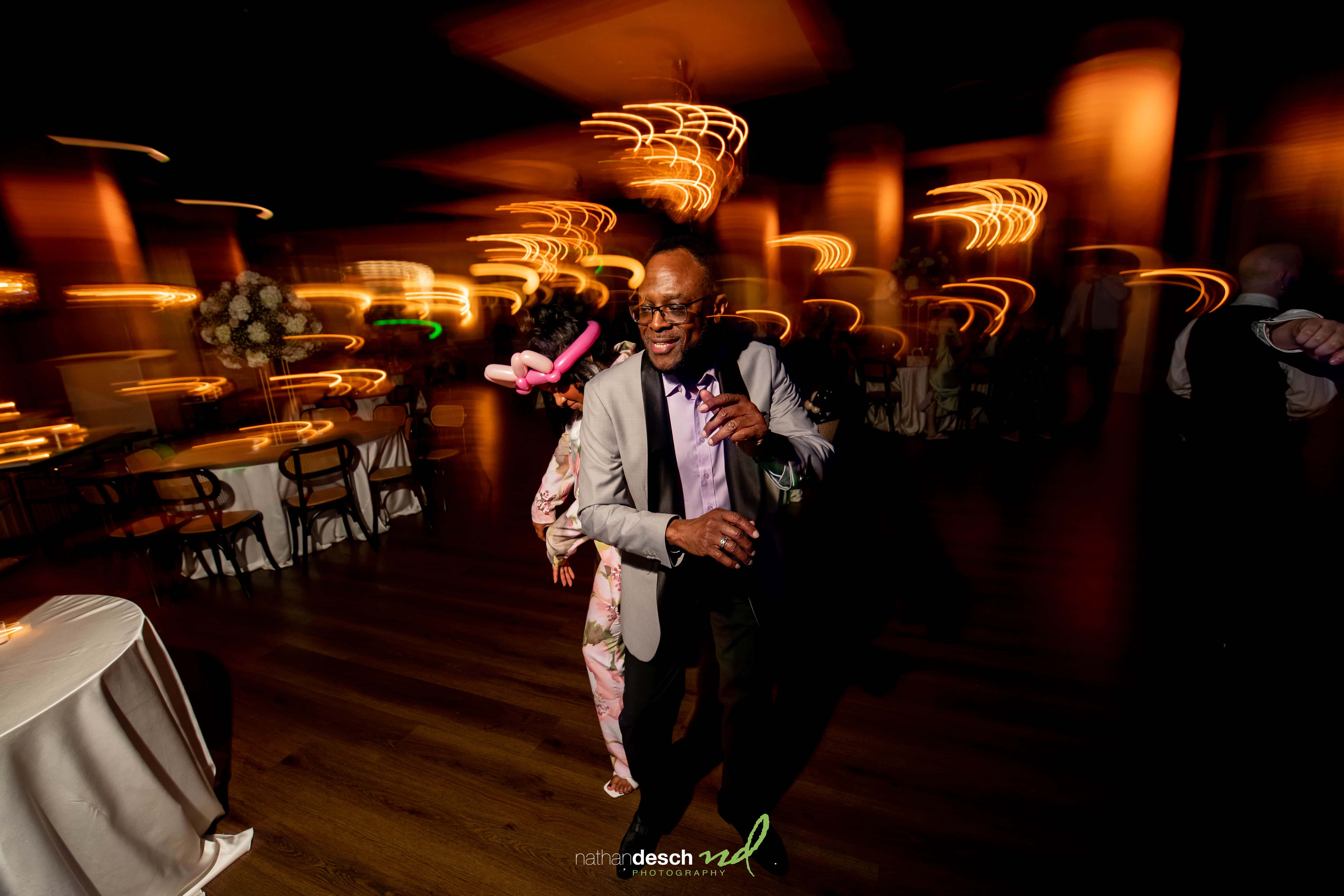 Joyful older man dancing under colorful lights at Tendenza spring wedding, surrounded by tables and guests.
