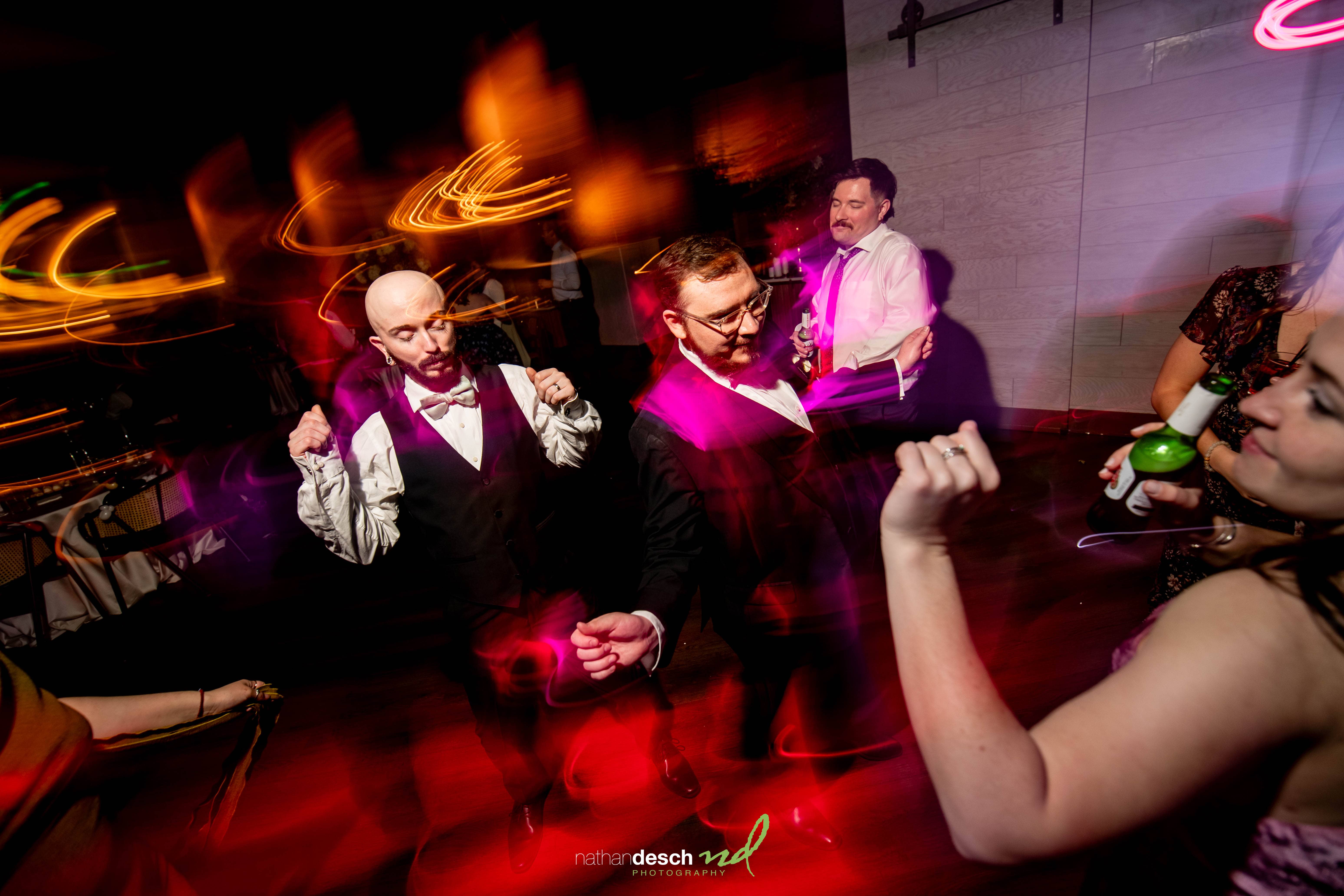 Two grooms dancing energetically at a vibrant spring Tendenza wedding, surrounded by colorful lights and joyful guests.