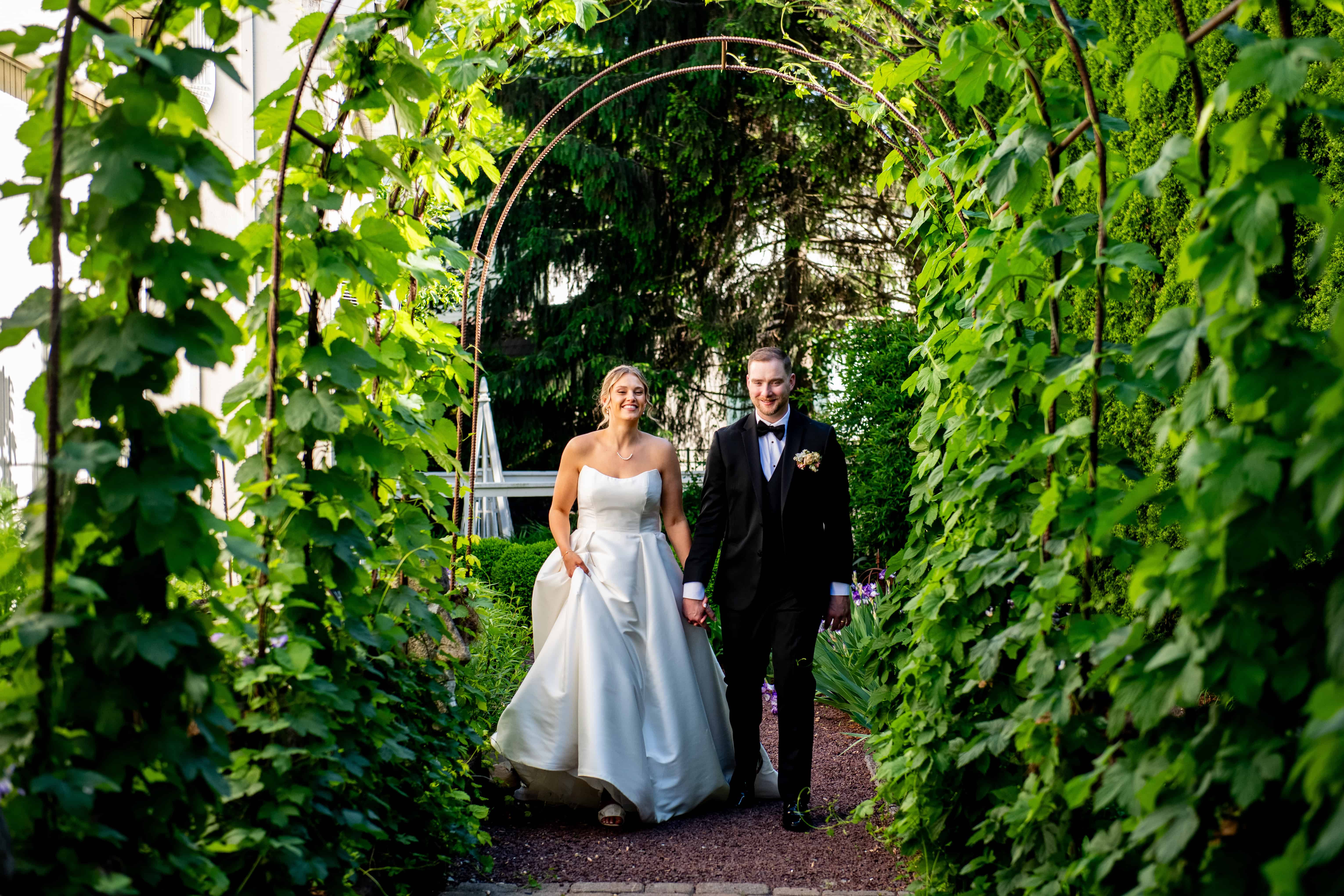 Bride and groom walking hand in hand through a lush garden archway at the Inn at Leola Village on their wedding day.