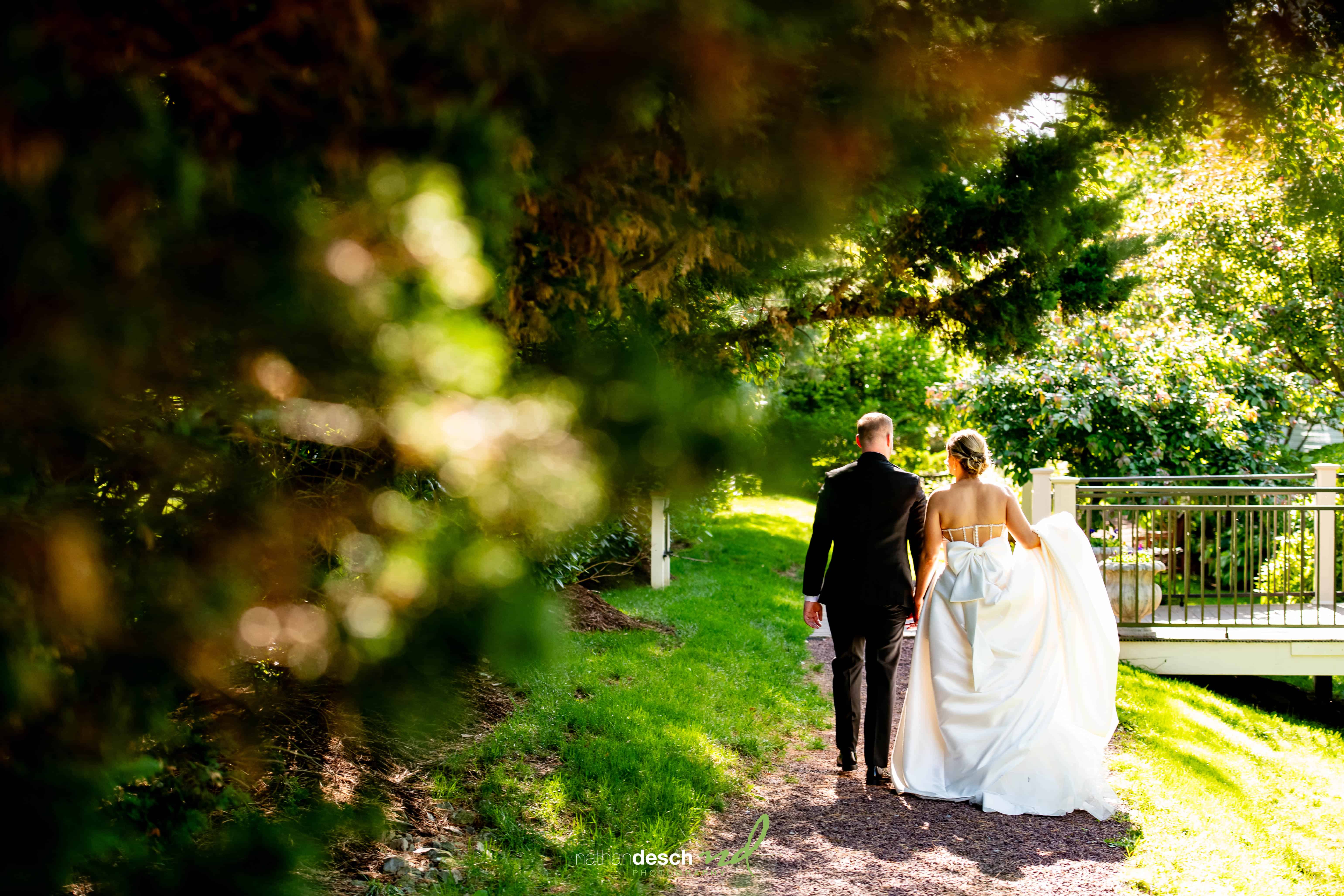 Bride and groom walk hand in hand through sunlit garden at The Inn at Leola Village, elegant wedding photography moment.