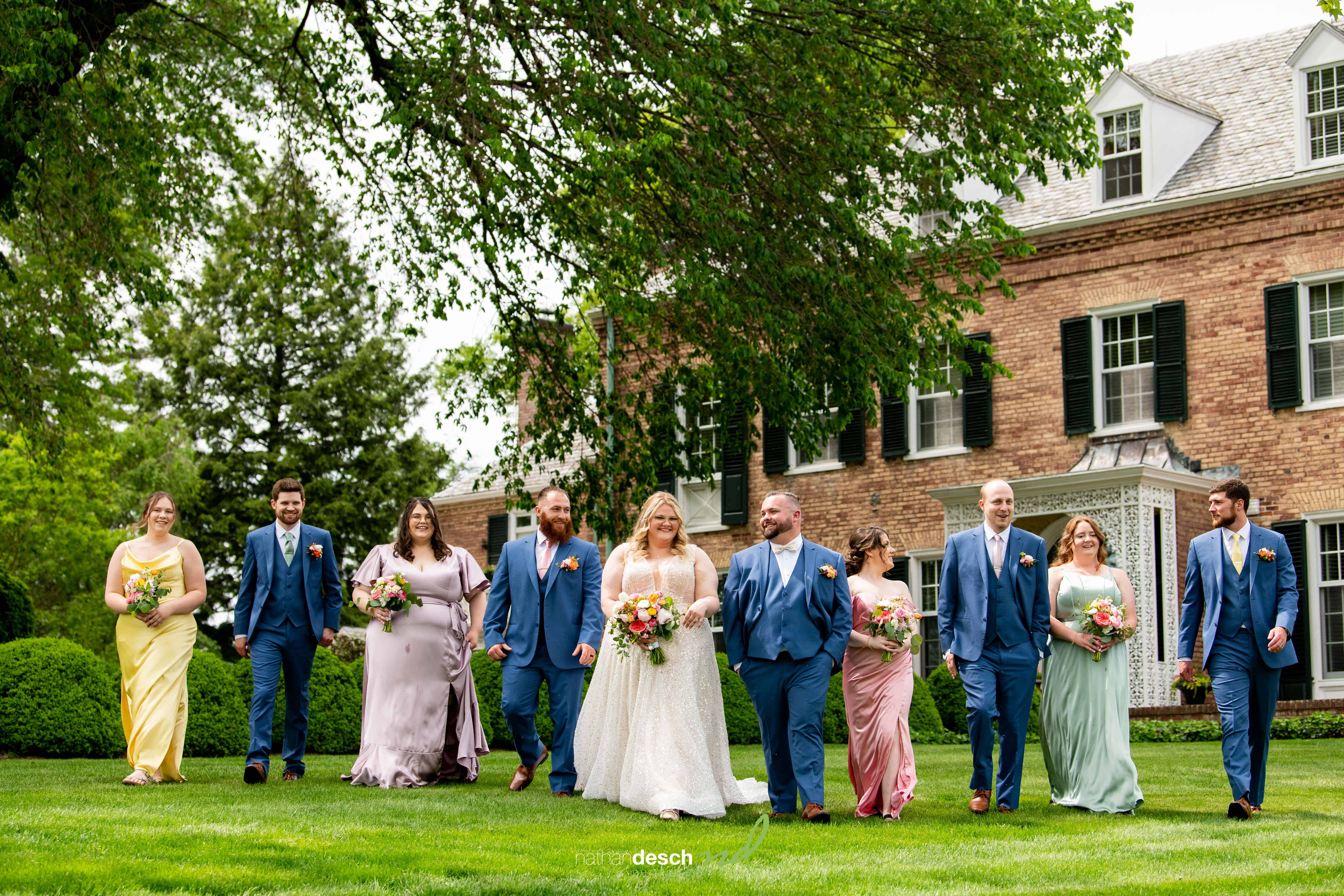 Wedding party of bridesmaids in colorful dresses and groomsmen in blue suits smiling at Drumore Estate with bouquets outdoors.