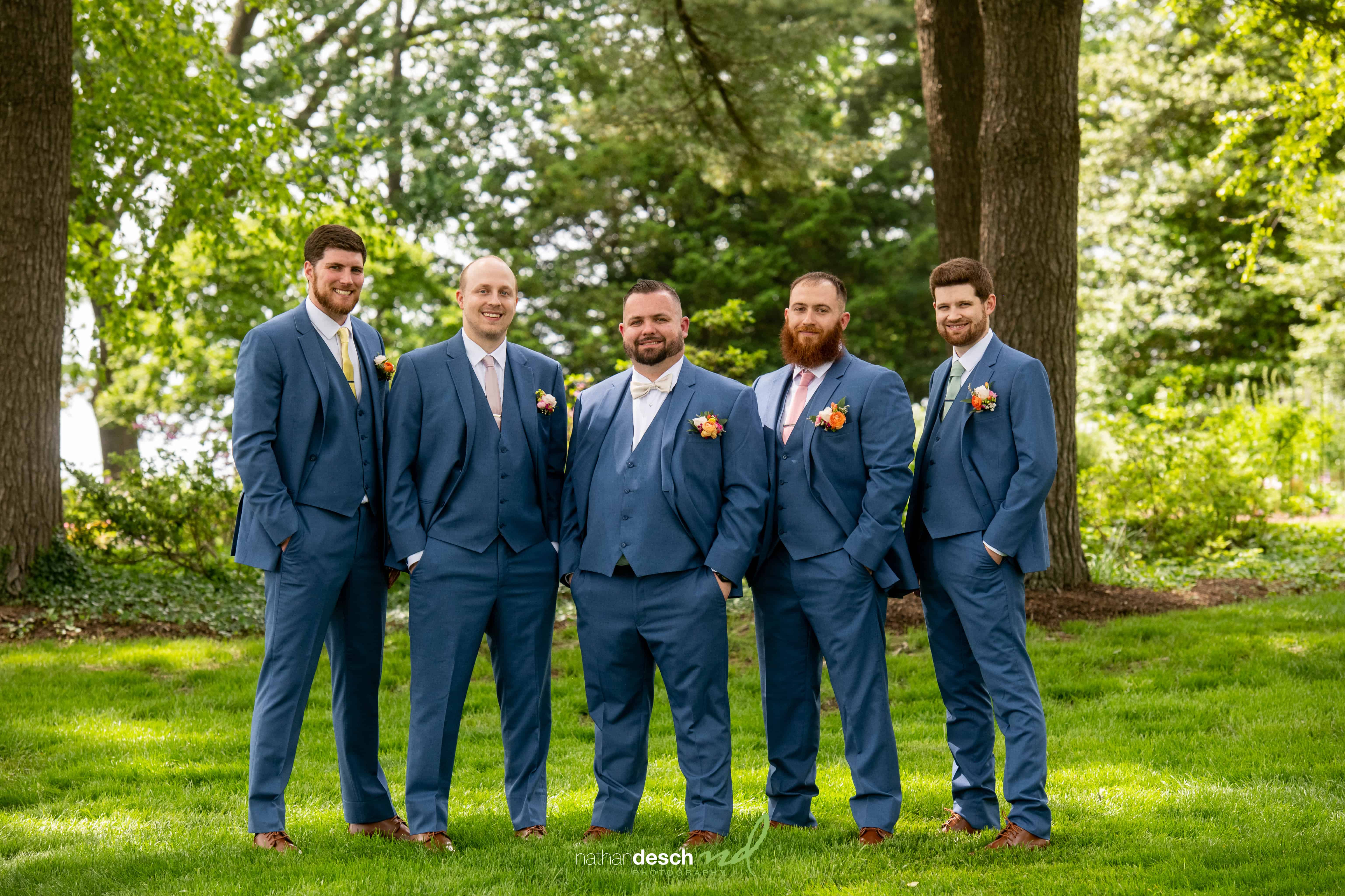 Groomsmen in blue suits with pink ties and orange boutonnieres smile outdoors at Drumore Estate spring wedding.