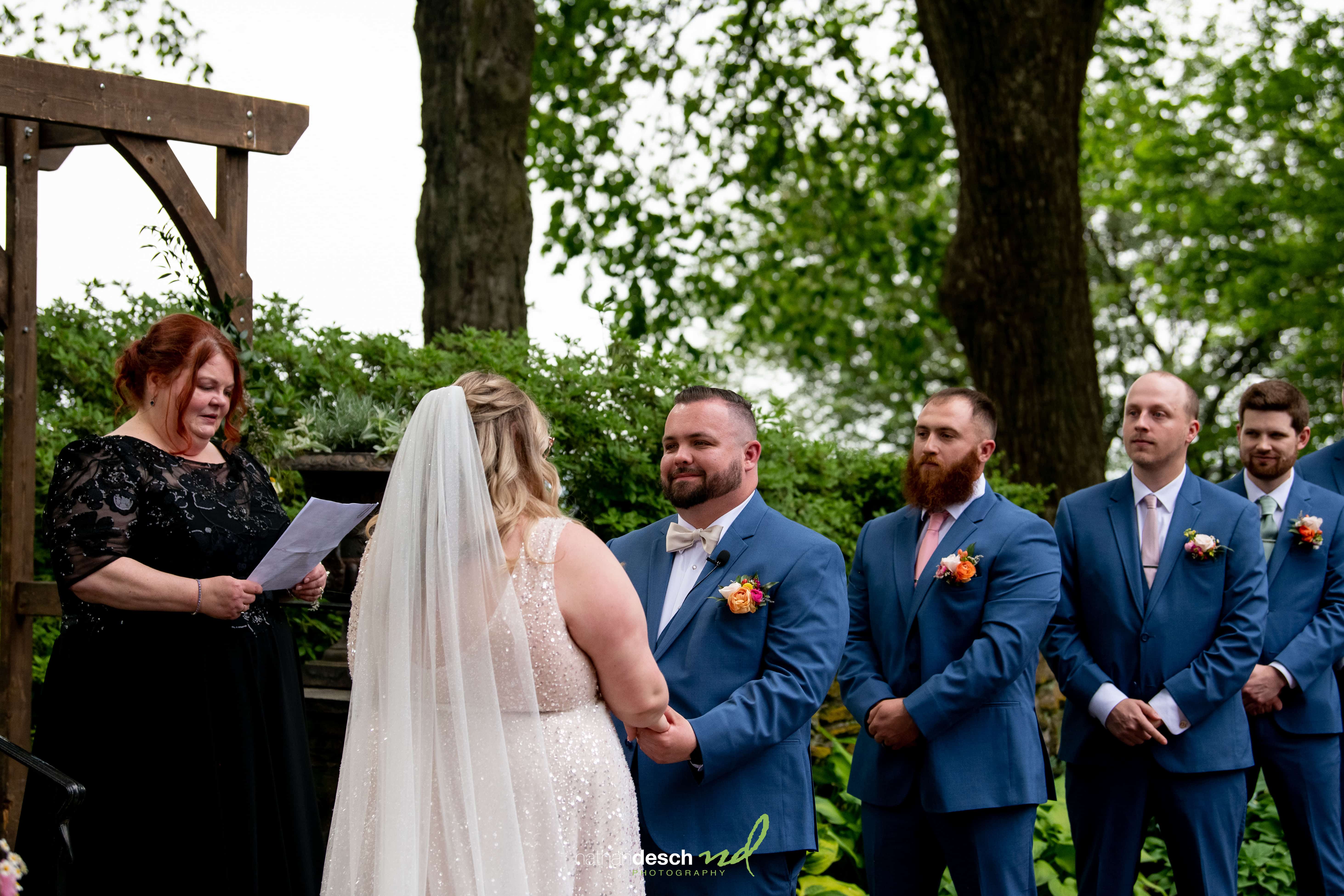 Bride and groom holding hands during outdoor Drumore Estate wedding ceremony with groomsmen in blue suits and lush greenery.