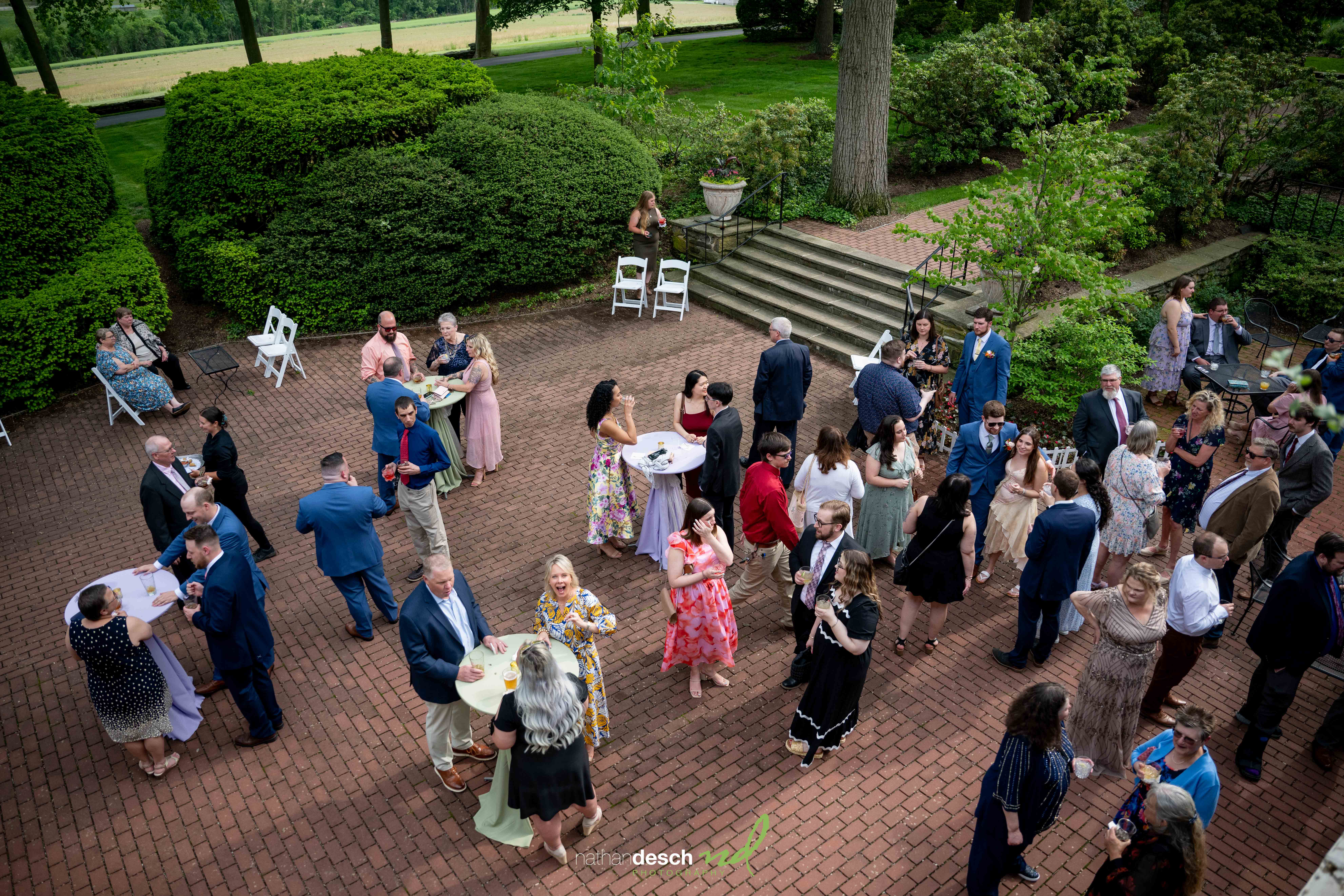 Wedding guests in elegant attire mingle on Drumore Estate’s brick patio, surrounded by spring garden greenery.