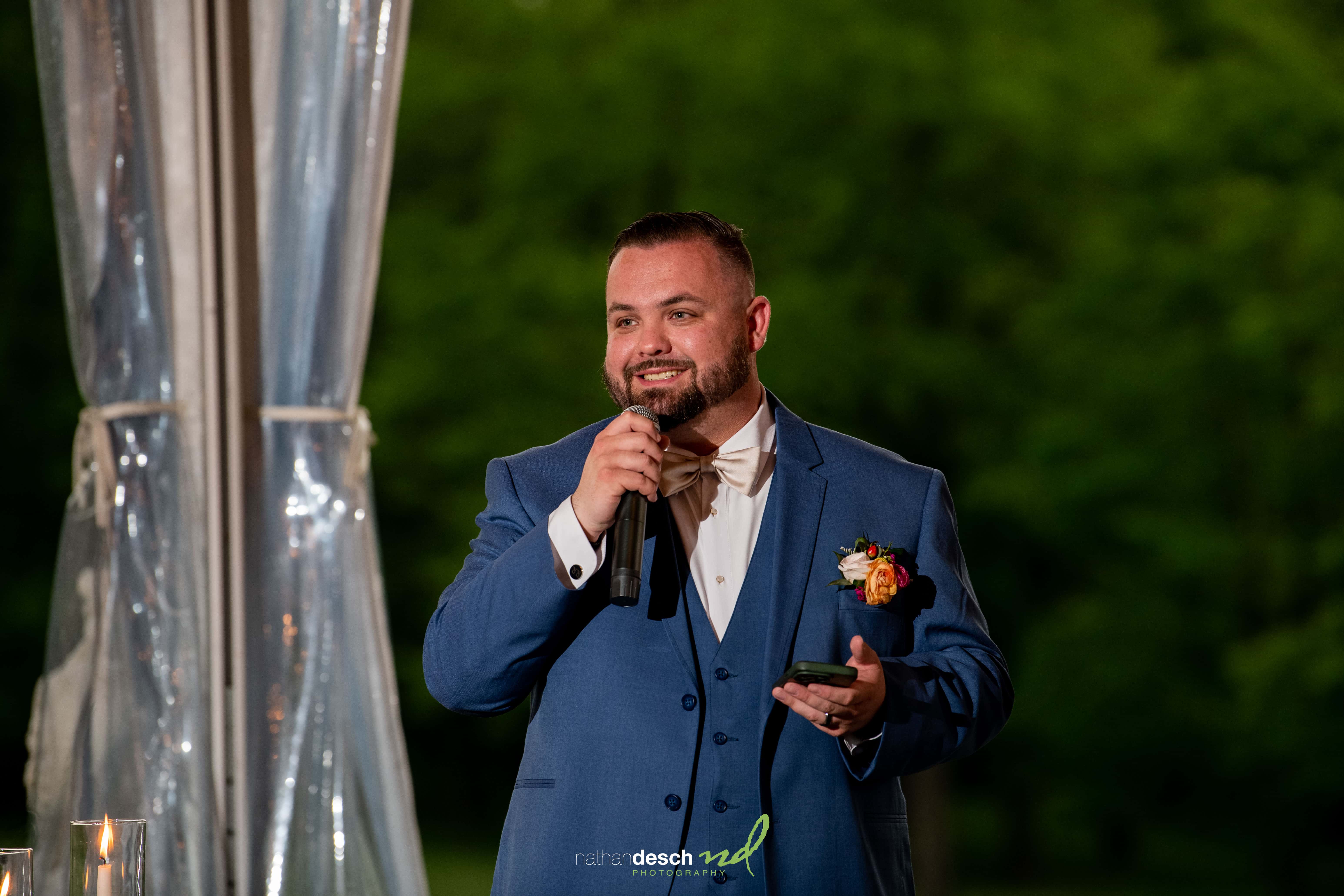 Man in blue suit giving wedding speech at Drumore Estate, with microphone, near elegant sheer curtains and lush greenery.
