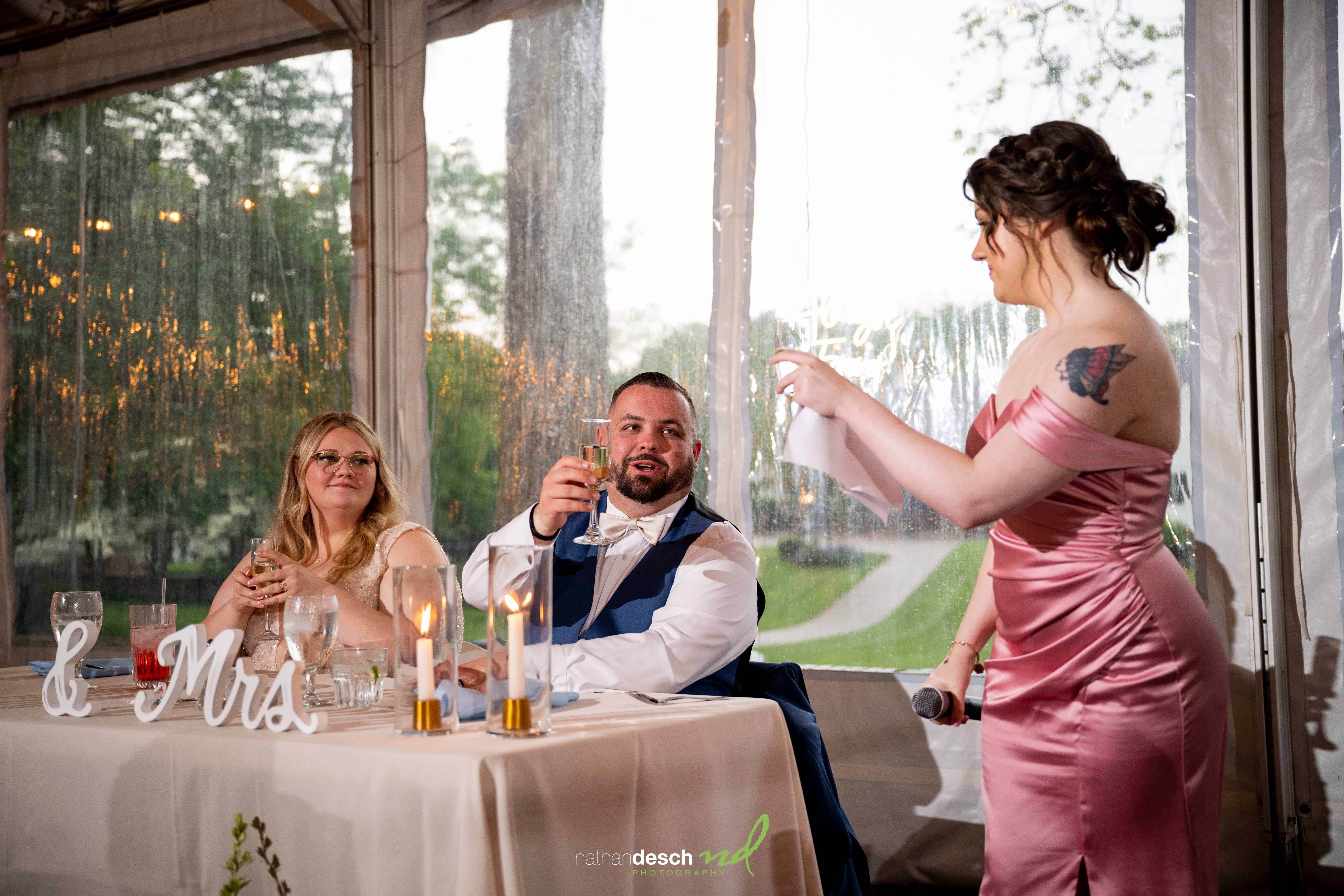 Woman in pink dress gives joyful wedding speech as newlyweds toast under clear tent at rainy spring Drumore Estate reception.